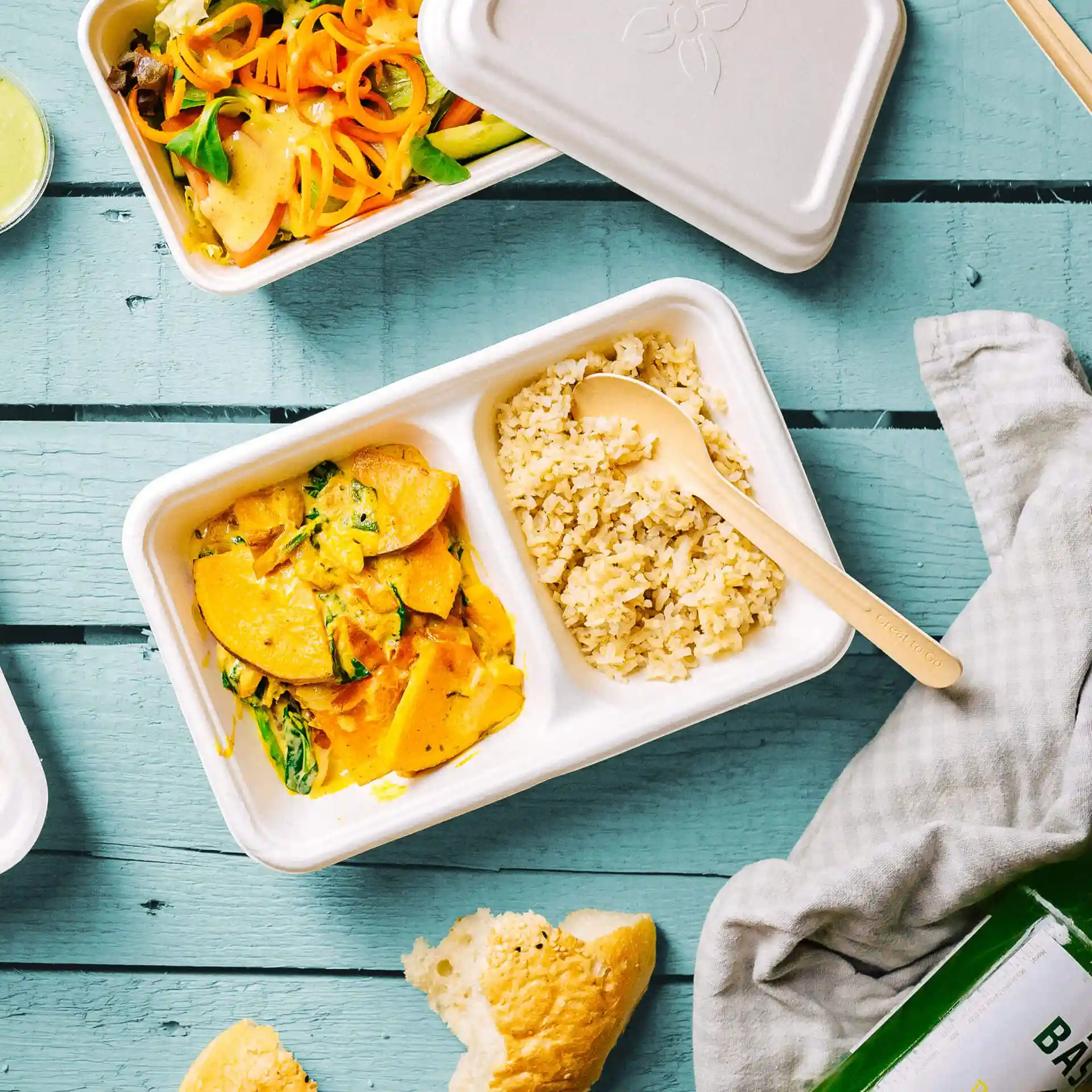 Rectangular white sugarcane tray with two compartments, containing rice and a vegetable dish, with a wooden spoon.