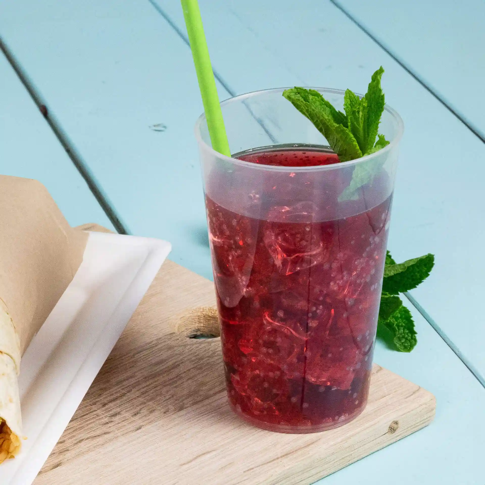 Transparent reusable cup filled with red drink and ice, with a green straw and mint leaves, placed on a wooden surface.
