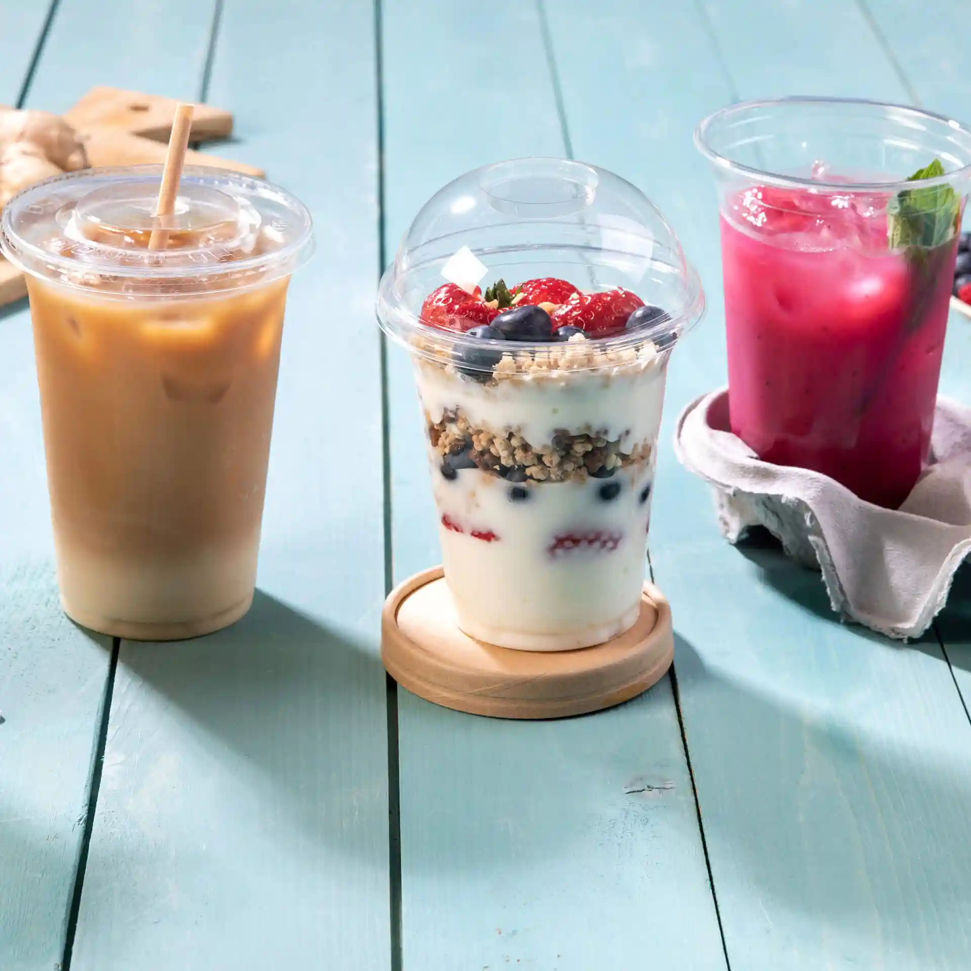 Bild von drei Getränken: Eiskaffee, Joghurtparfait mit Beeren und einem pinken Getränk auf einer blauen Holzoberfläche.