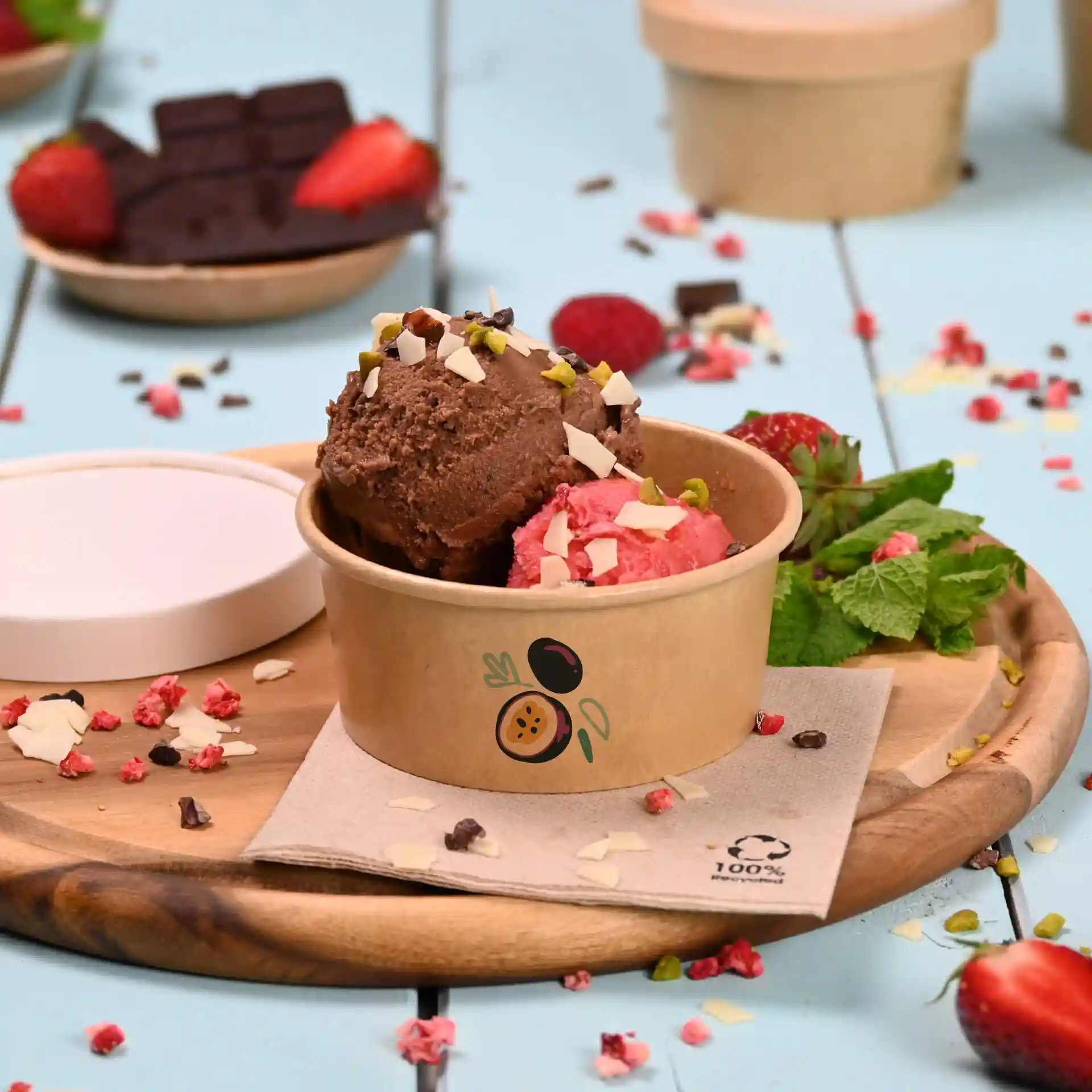 Brown cardboard ice cream cup with two scoops of chocolate and strawberry ice cream, topped with nuts and chocolate pieces.