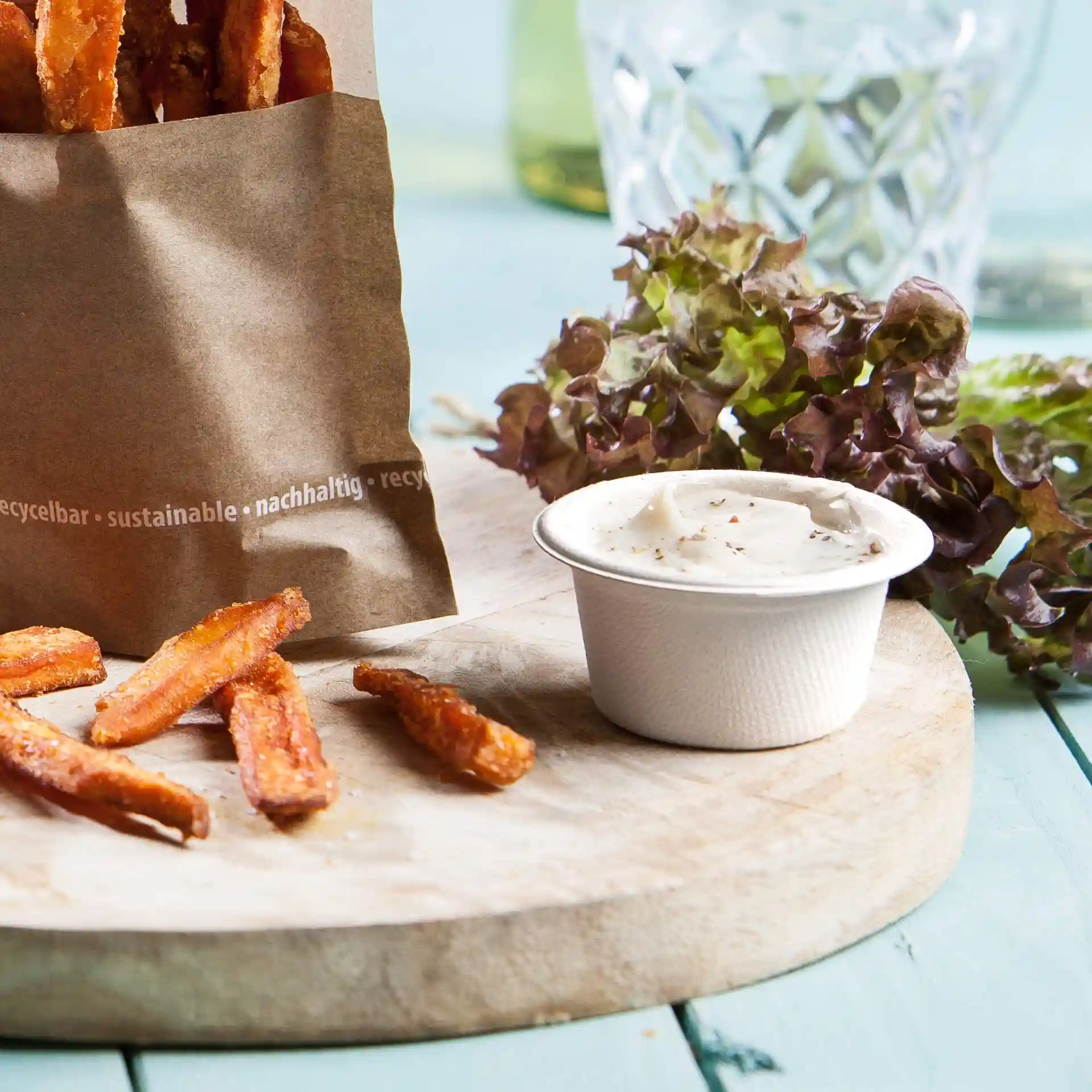 Sweet potato fries in a paper bag, with a small cup of dip and fresh lettuce on a wooden board.