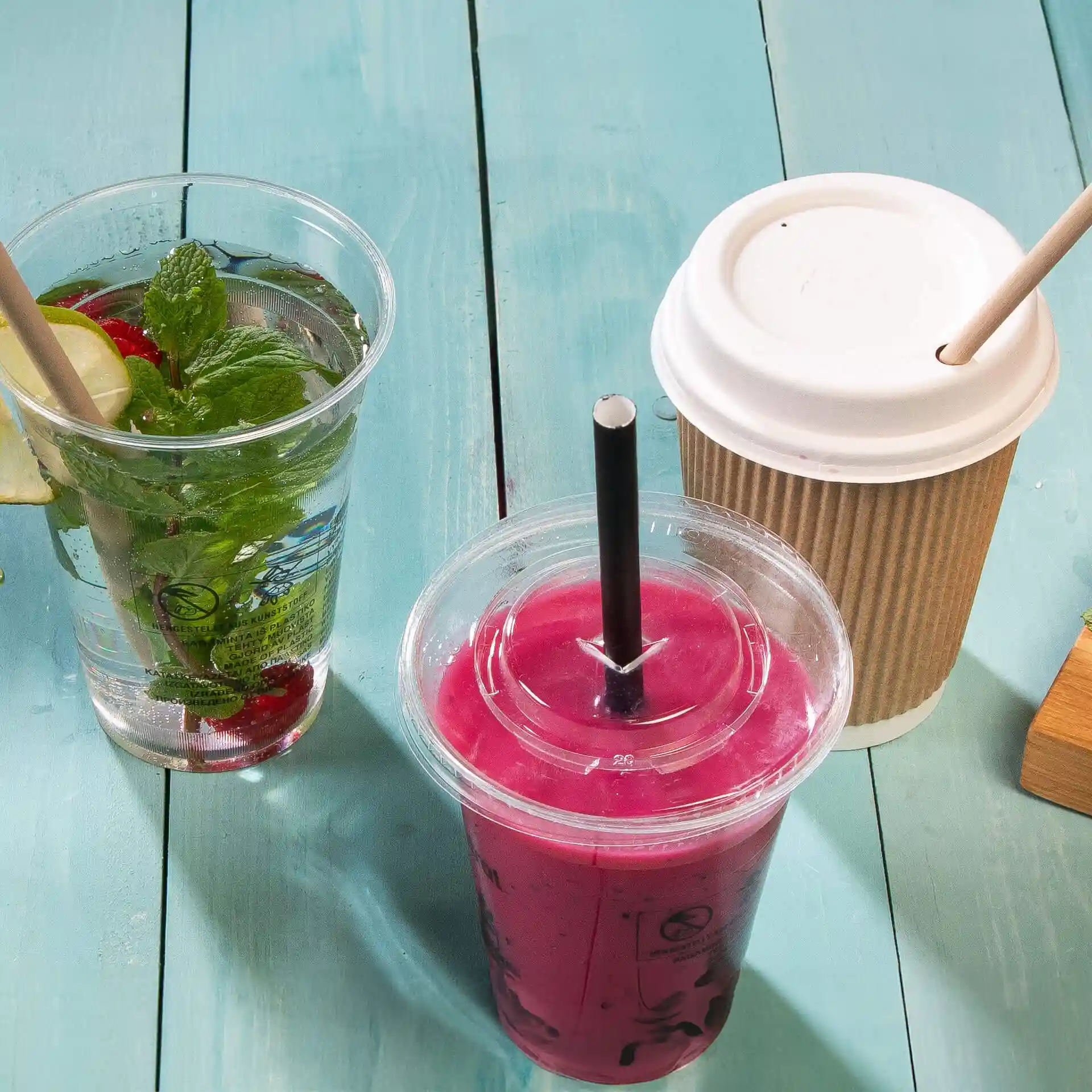 Image of three drinks on a blue wooden surface: a clear cup with mint and lemon, a pink drink with a black straw, and a paper cup.