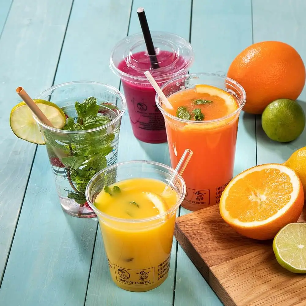 Vier klare Plast Becher gefüllt mit bunten Getränken, darunter Orangensaft und ein rosa Smoothie, neben frischen Früchten.