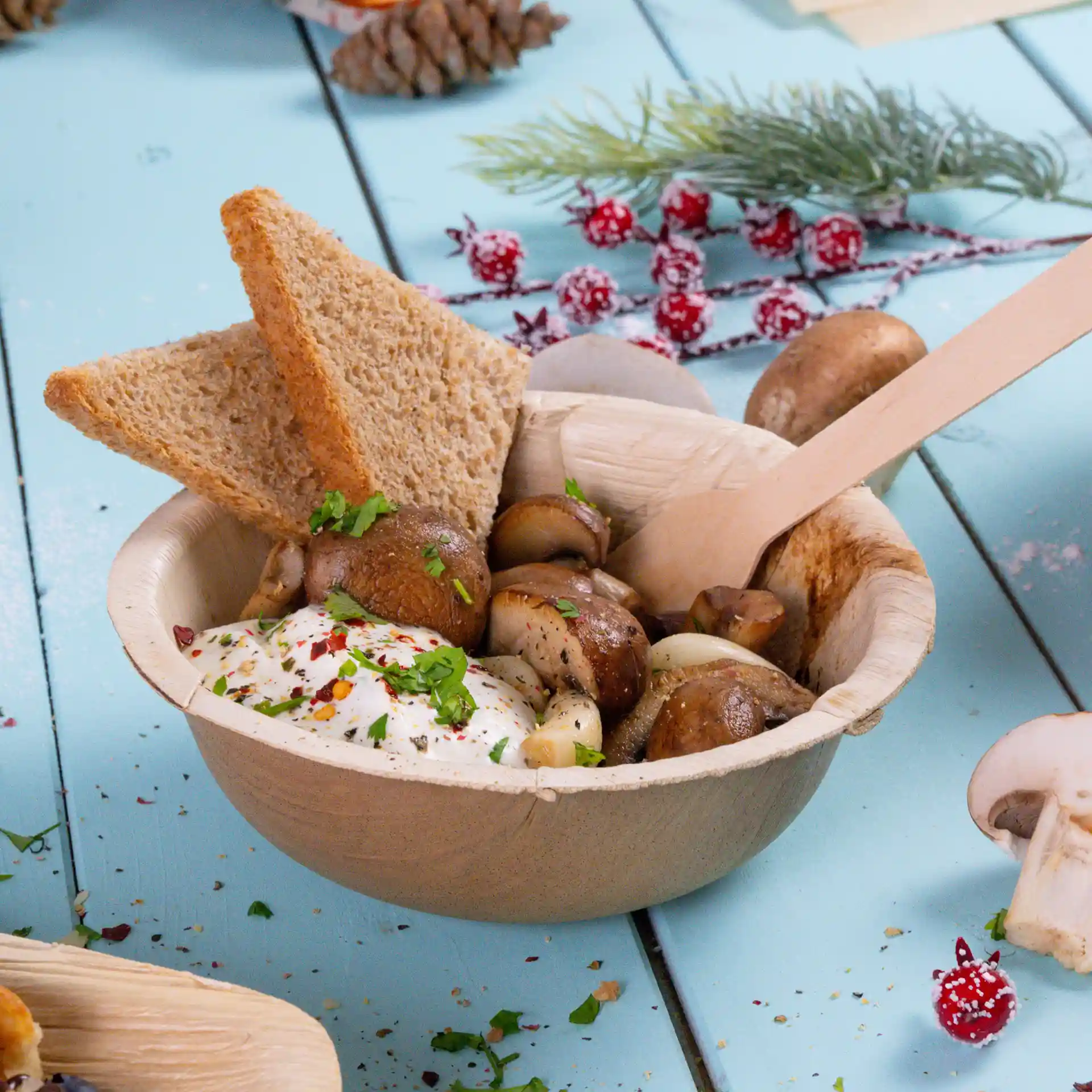 Schüssel aus Palmblatt mit Pilzen, einem Klecks Sauce und zwei Scheiben Vollkornbrot, mit einem Holzlöffel.