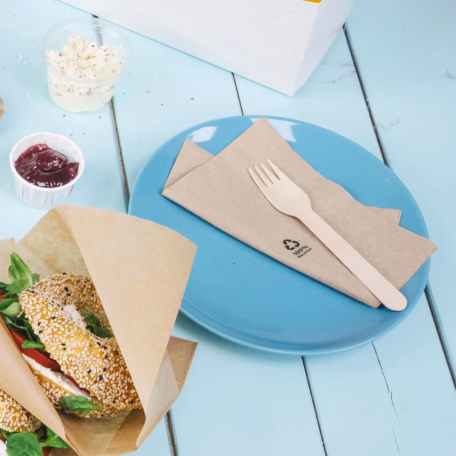 Ein blauer Teller mit gefalteten braunen Servietten und einer Holzgabel; ein Sesam-Bagel mit Salat daneben, Marmelade in einem Becher.