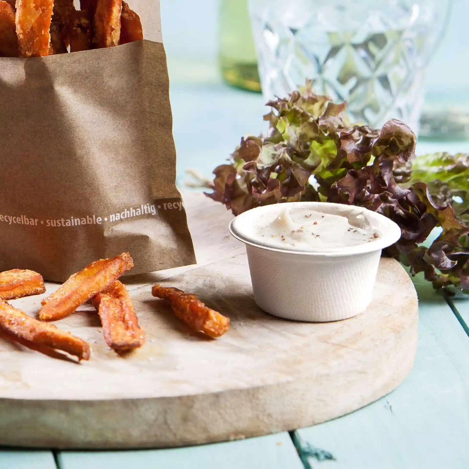 Image of a paper bag filled with sweet potato fries, a white dip bowl, and leafy greens on a wooden board.
