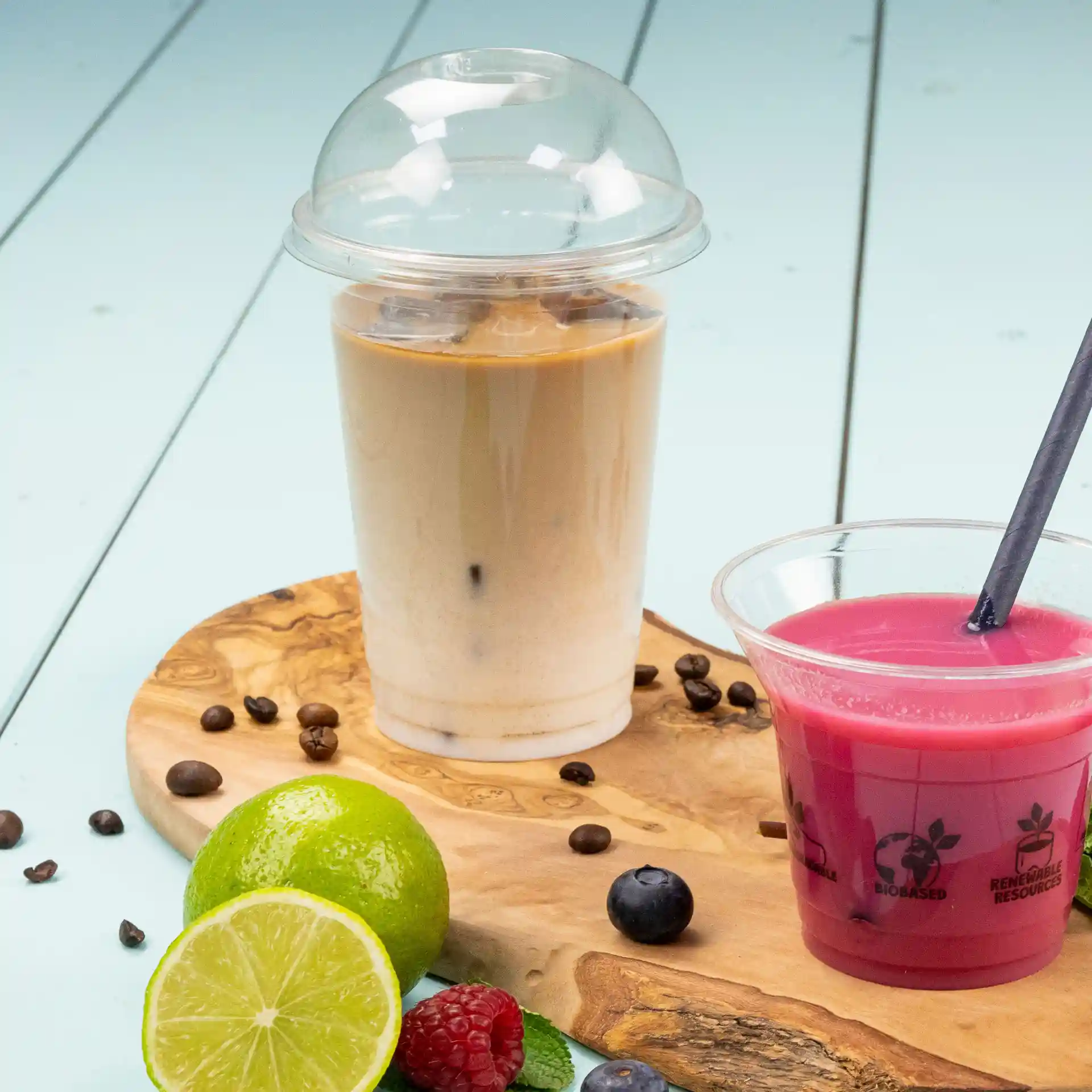Bild eines transparenten 16 oz Plastikbechers, gefüllt mit Eiskaffee, neben einem pinken Getränk in einem klaren Becher auf einem Holzbrett mit Früchten.