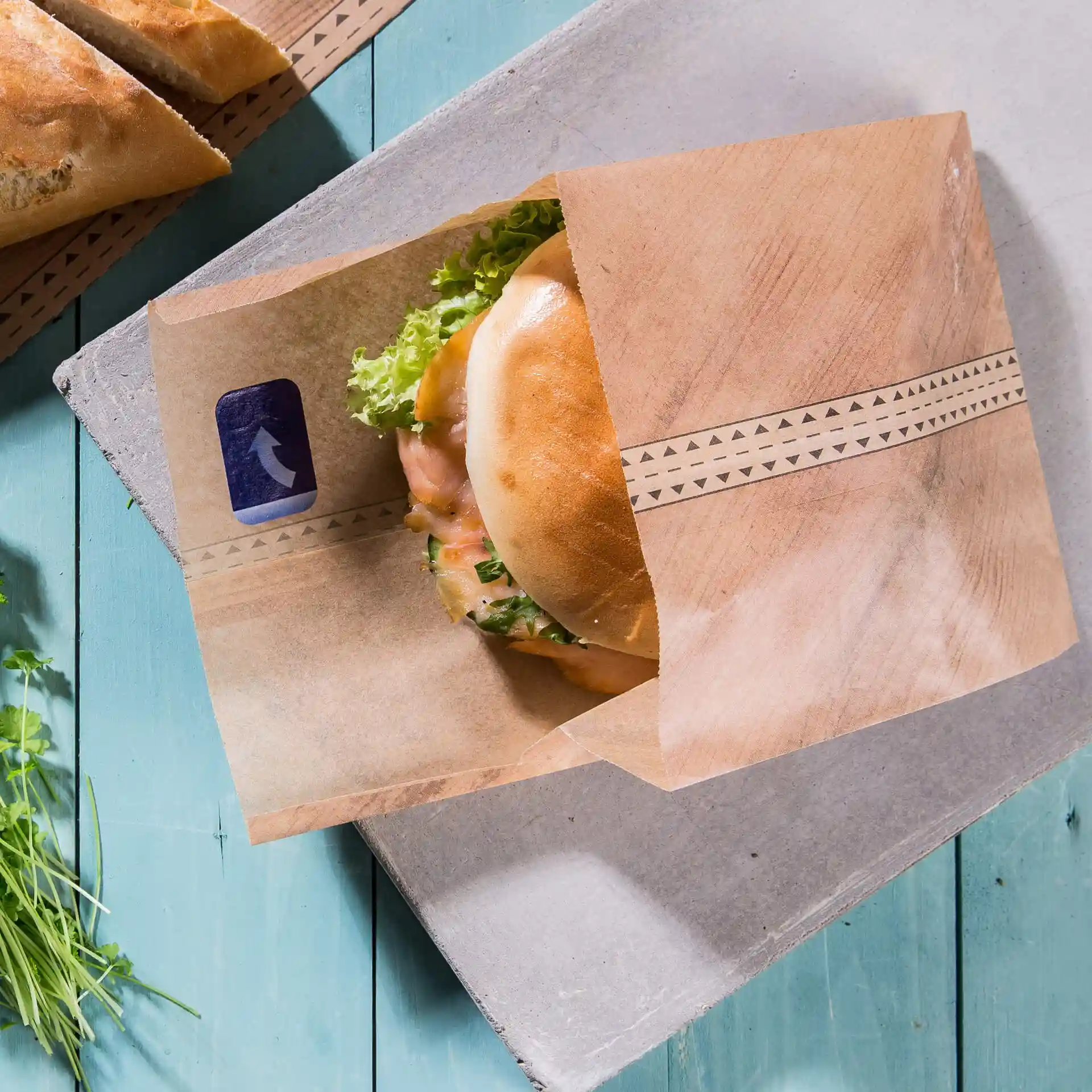 Braune Papiertüte mit einem Burger und Salat, auf einer blauen Holzoberfläche platziert.