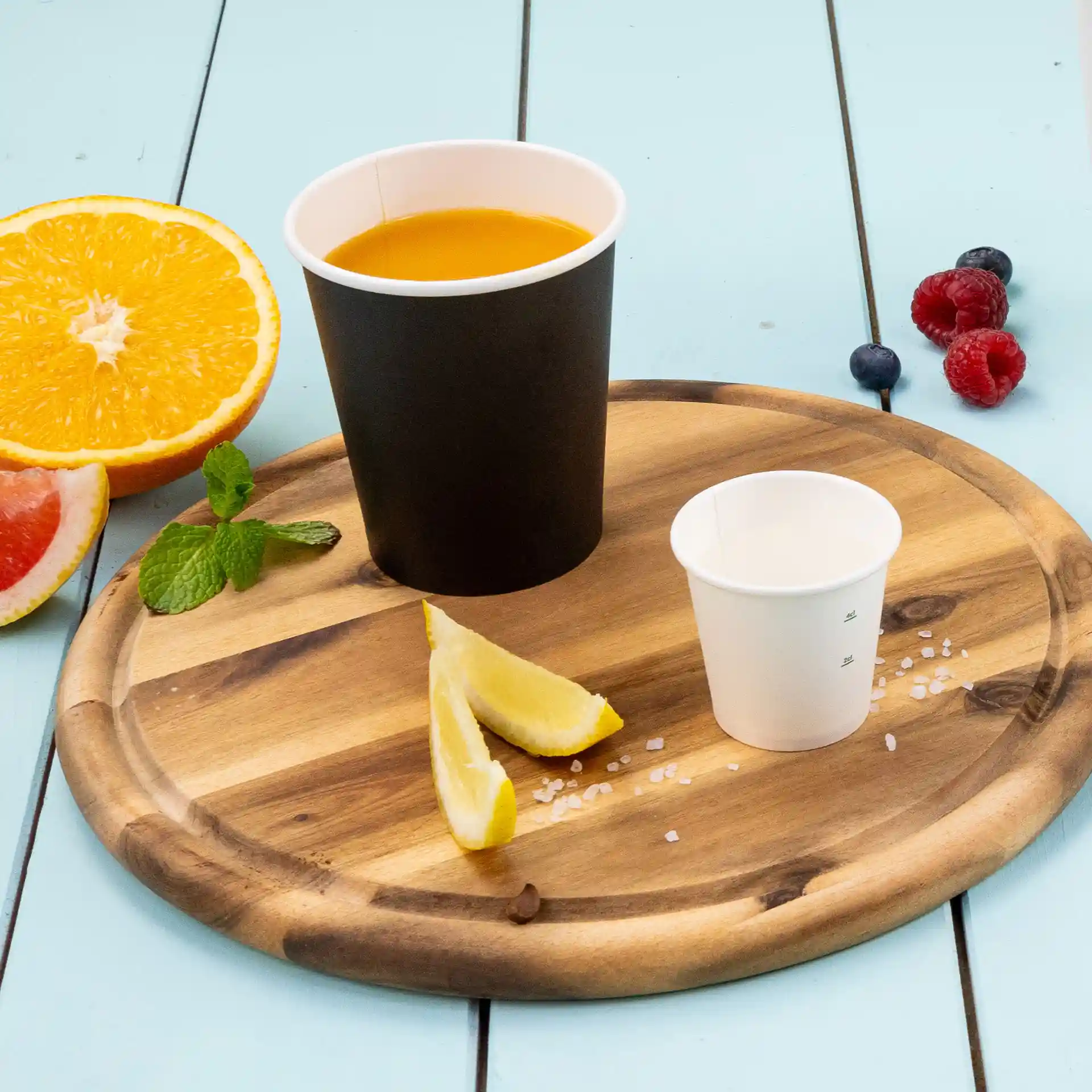 Black paper cup with orange liquid, small white cup, lemon wedges, and fresh fruit on a wooden tray.