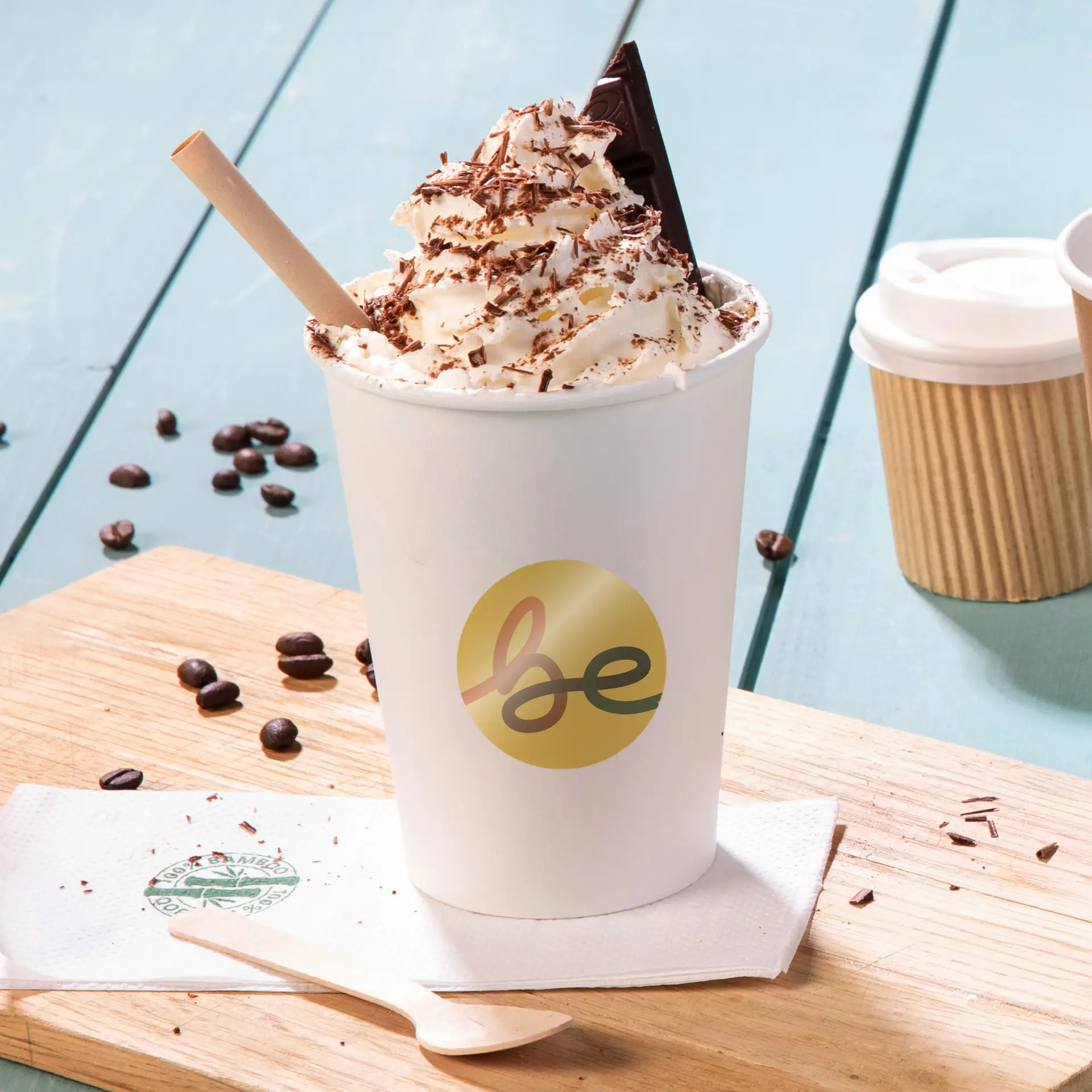 Cup with whipped cream topped with chocolate flakes, a chocolate piece, and a paper straw, sitting on a wooden surface with coffee beans.