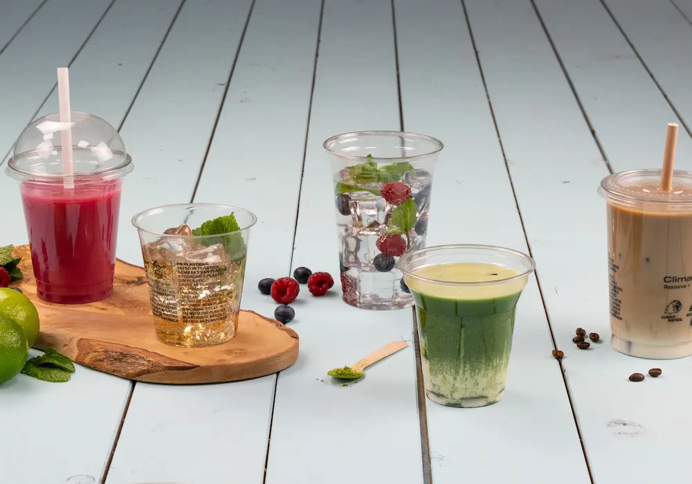 Five cups of different beverages on a wooden tray, featuring pink, green, clear, and brown drinks with fruits and mint.