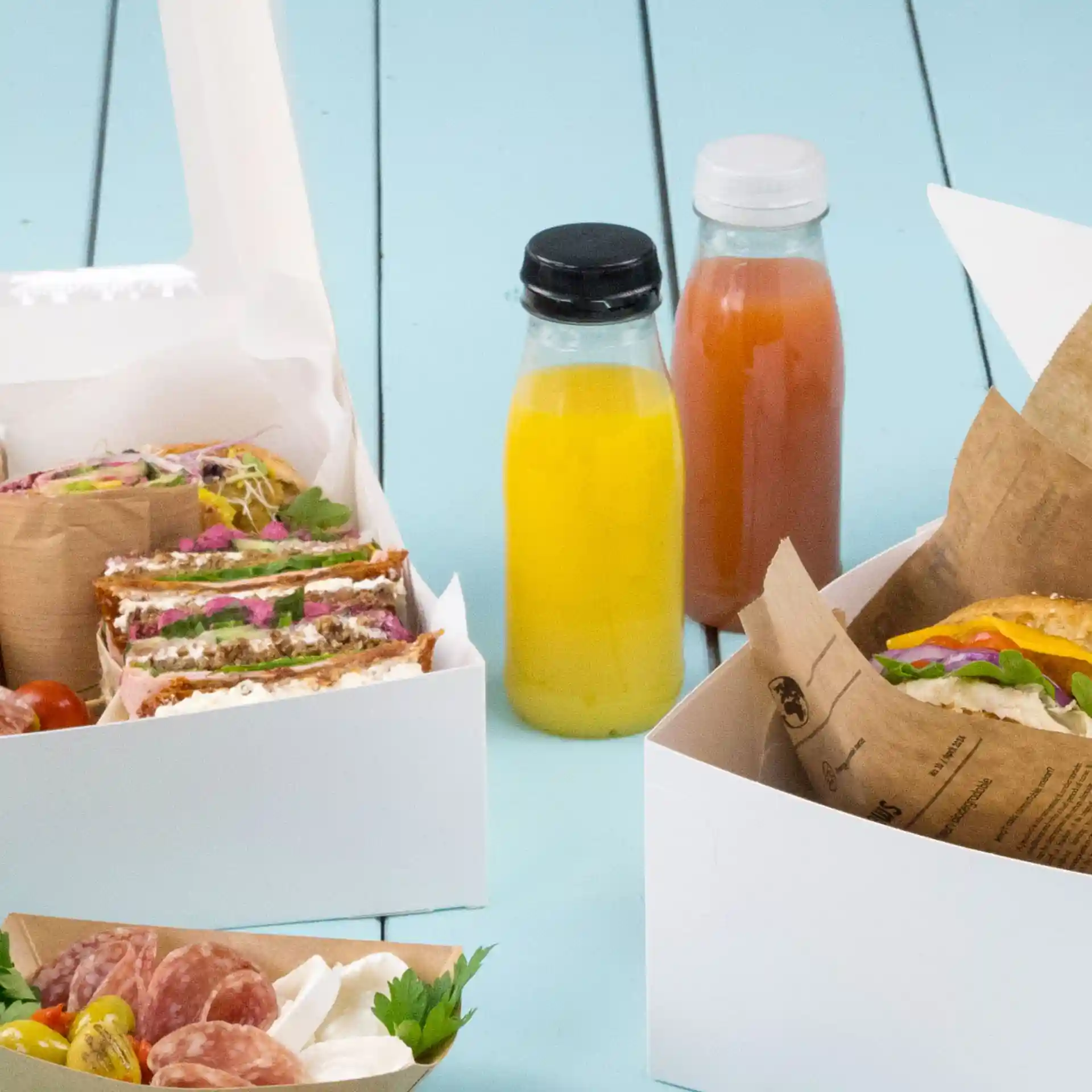 Bild von Lebensmittelbehältern mit Sandwiches, einem Salat und zwei klaren Flaschen mit schwarzen und weißen Deckeln auf einer blauen Oberfläche.