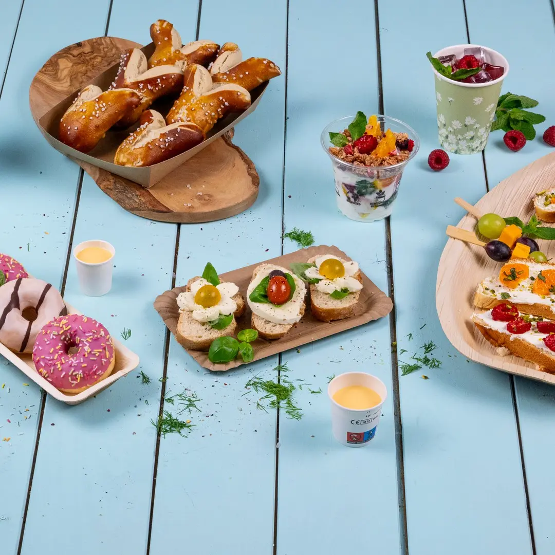 Eine Auswahl an Fingerfood, darunter Brezeln, belegte Sandwiches, Donuts und Obstbecher auf einem blauen Holztisch.
