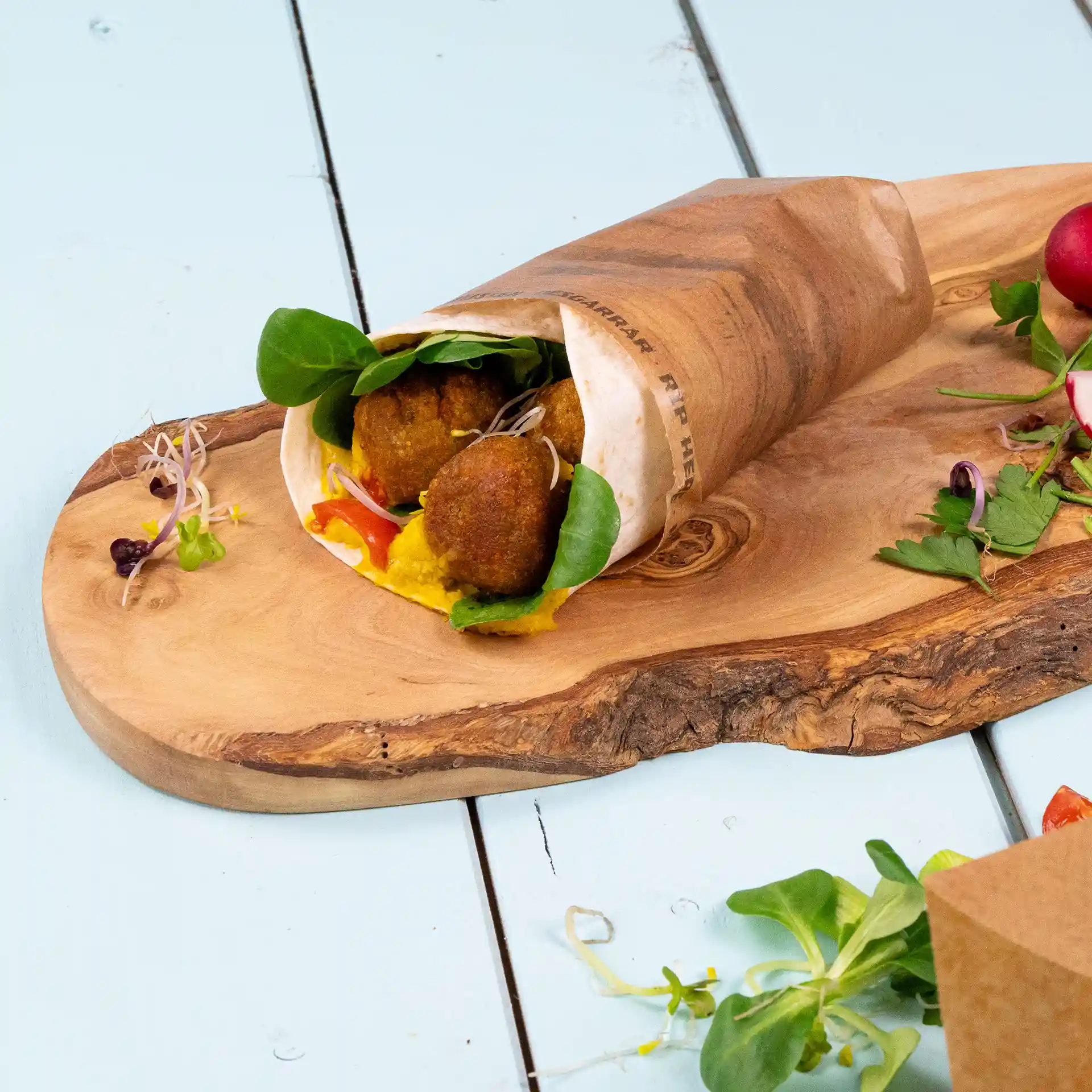Braune Kraft-Burritotasche mit Falafel, Gemüse und Sauce, auf einem Holzbrett mit zusätzlichem Gemüse daneben.
