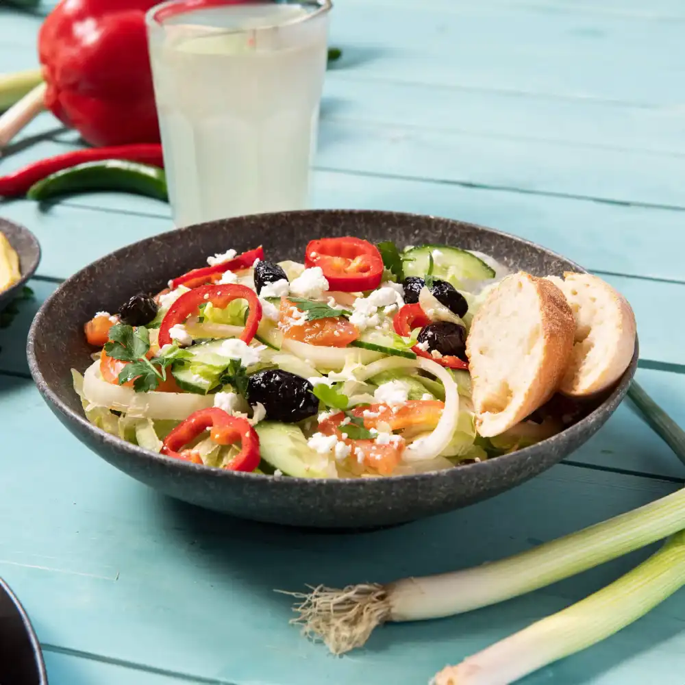 Schüssel mit Salat, Gurken, Tomaten, Oliven, Feta und roten Paprikastücken, dazu zwei Brote.