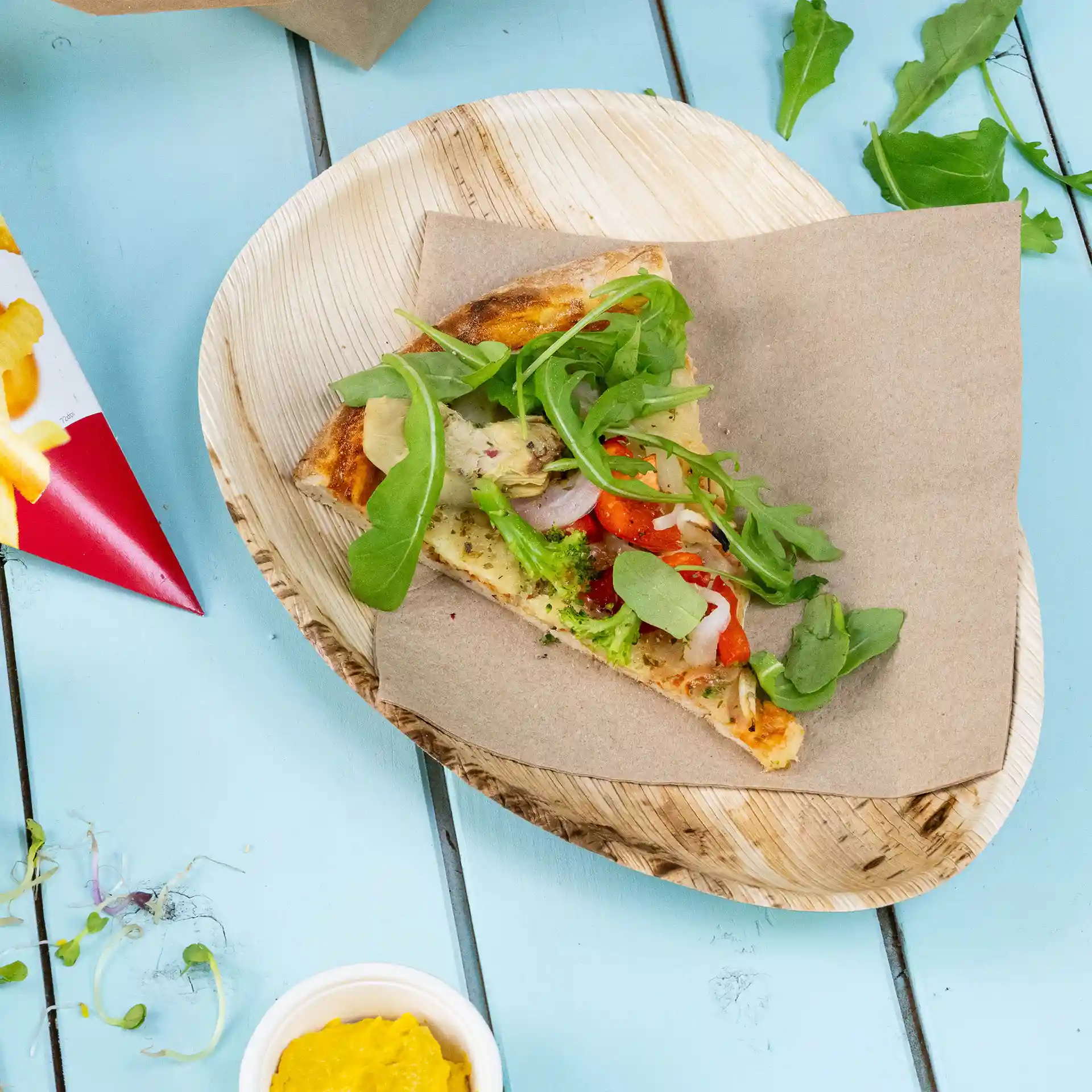 Stück Pizza mit Gemüse auf braunem Papier auf einem Palmblatt-Teller auf einer blauen Oberfläche.