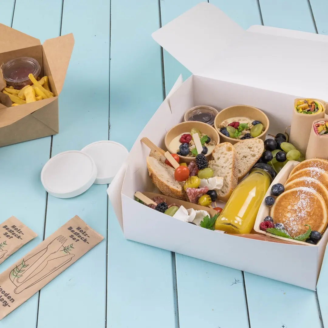 Weißer Take-away-Behälter gefüllt mit verschiedenen Lebensmitteln, darunter Brot, Obst, Pfannkuchen und ein Getränk, auf einer blauen Holzoberfläche.