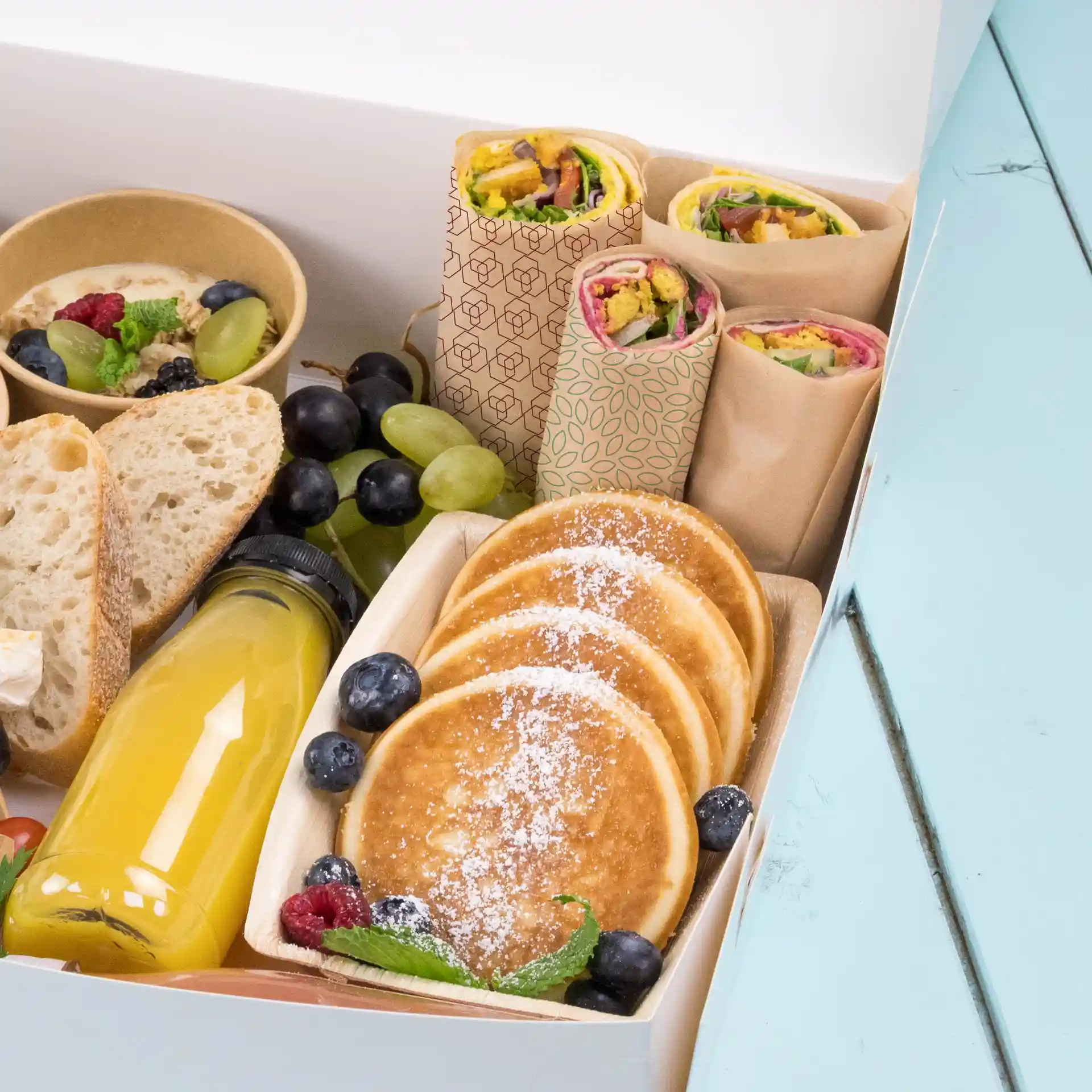 Verschiedene Lebensmittel wie Wraps, Pfannkuchen, Obst und ein Getränk in einer Box auf einer blauen Oberfläche.