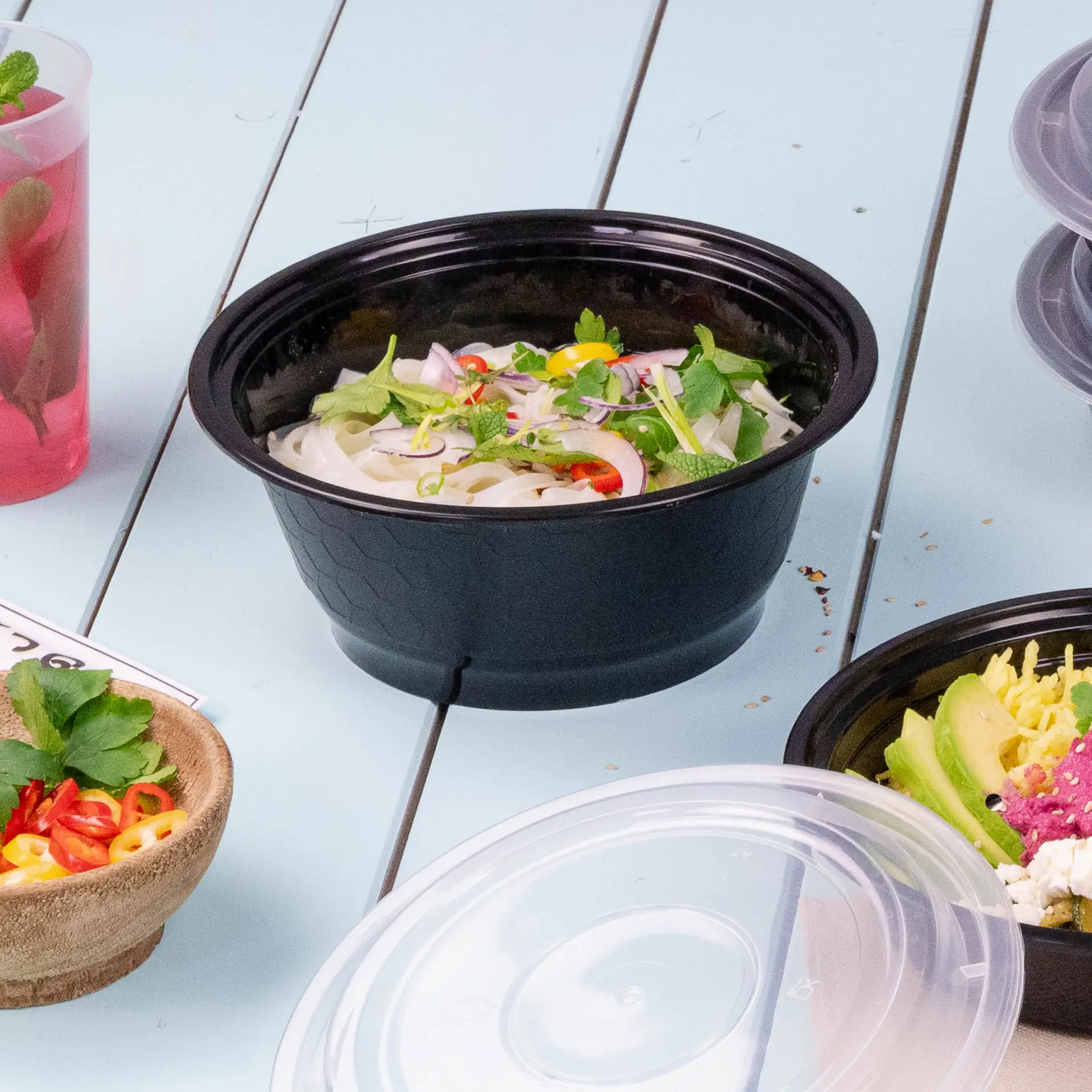 One black reusable container filled with noodles and garnished with fresh herbs and chili; other food items are nearby.