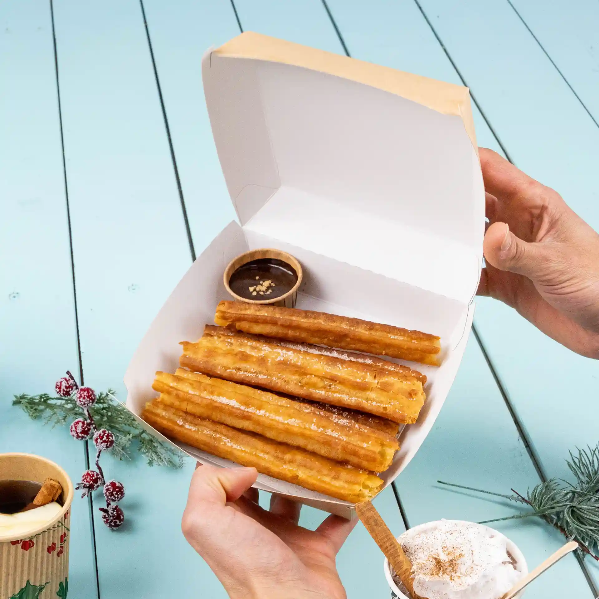 Takeaway box holding several churros with a small cup of chocolate sauce, on a light blue wooden surface.