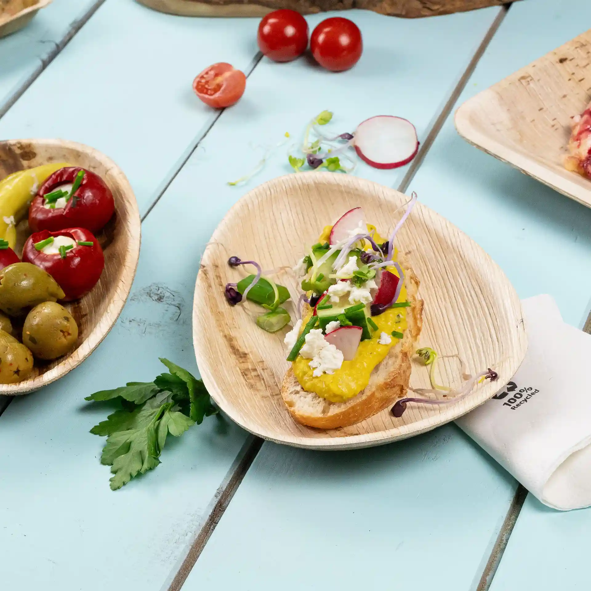 Bild eines flachen ovalen Palmblatt-Tellers mit einer Scheibe Brot, belegt mit gelber Paste, Gemüse und Kräutern, neben Oliven und Tomaten.