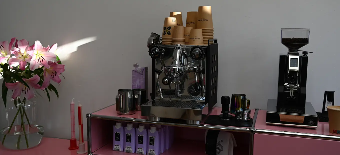 Kaffeemaschine mit gestapelten Bechern, Kaffeemühle und pinken Blumen auf einer Arbeitsplatte. Verschiedene kaffeemaschinenbezogene Gegenstände sind sichtbar.