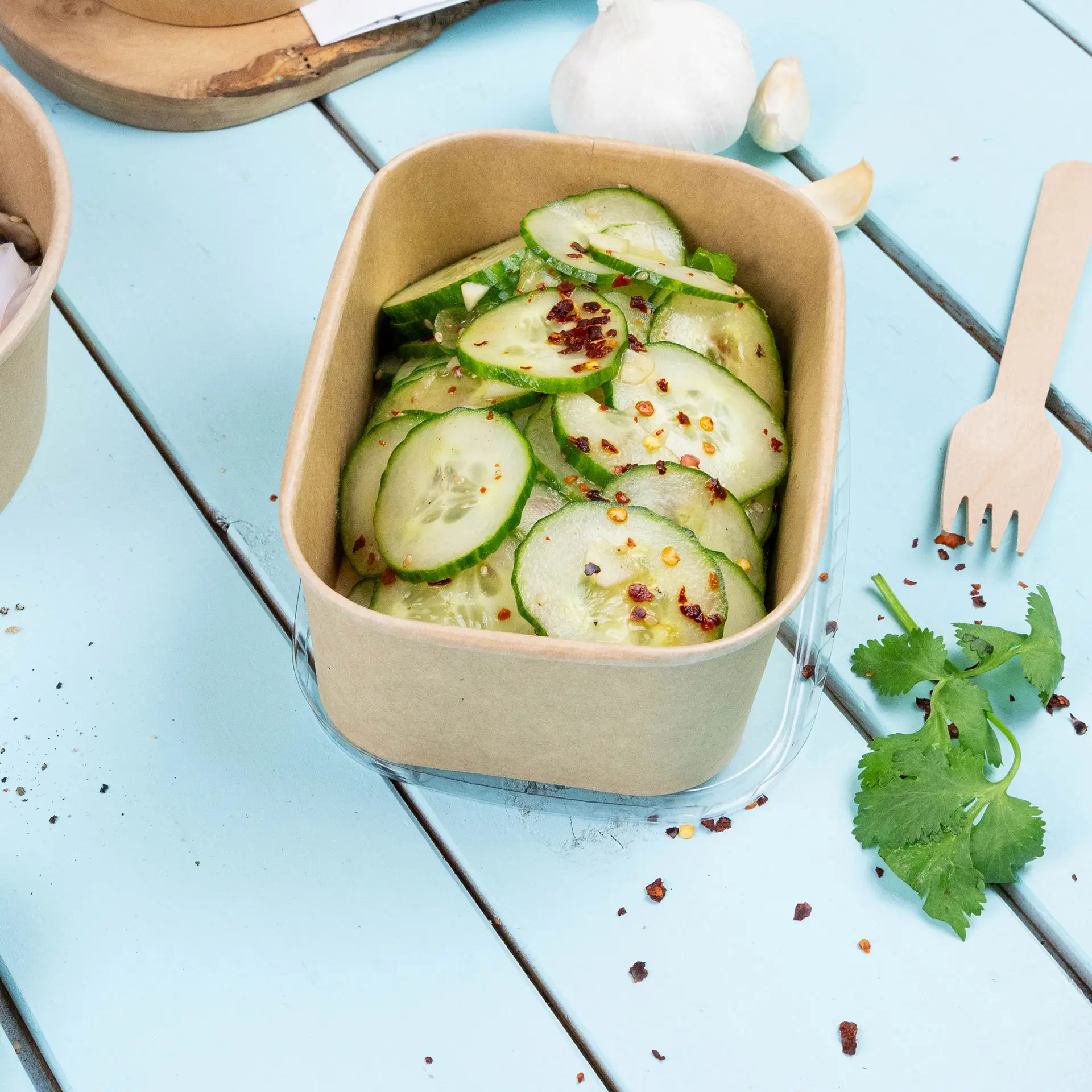Brauner Kraft-Lebensmittelbehälter gefüllt mit geschnittenen Gurken, mit Chiliflocken bestreut, auf einer hellblauen Holzoberfläche.