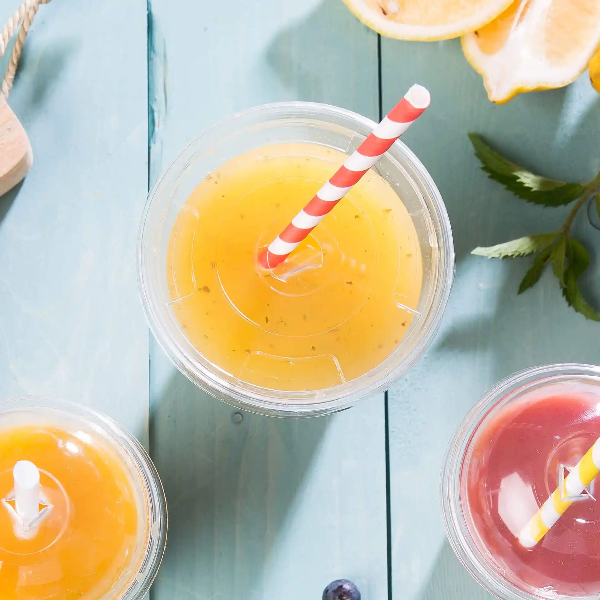 Transparent PLA flat lid with a straw hole, placed above colorful drinks on a blue wooden surface.
