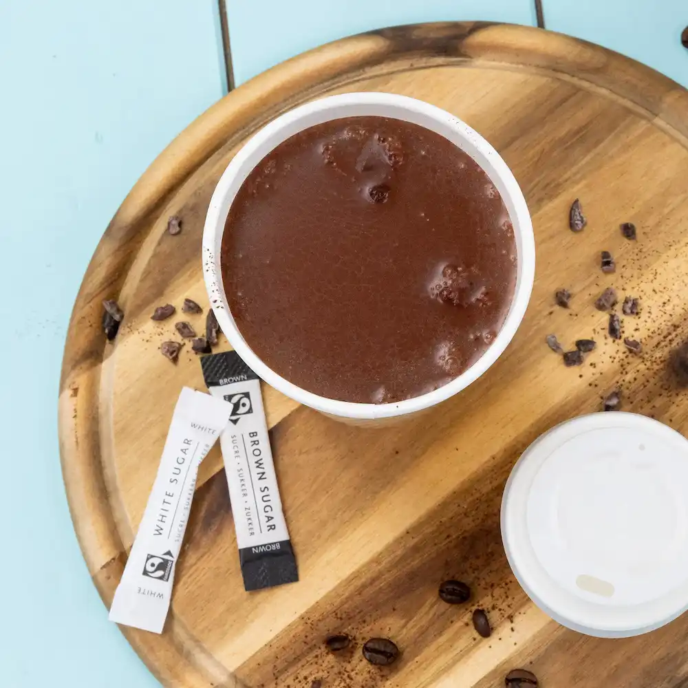 Hot chocolate in a white cup with sugar packets, on a wooden serving tray with coffee beans and spilled cocoa.