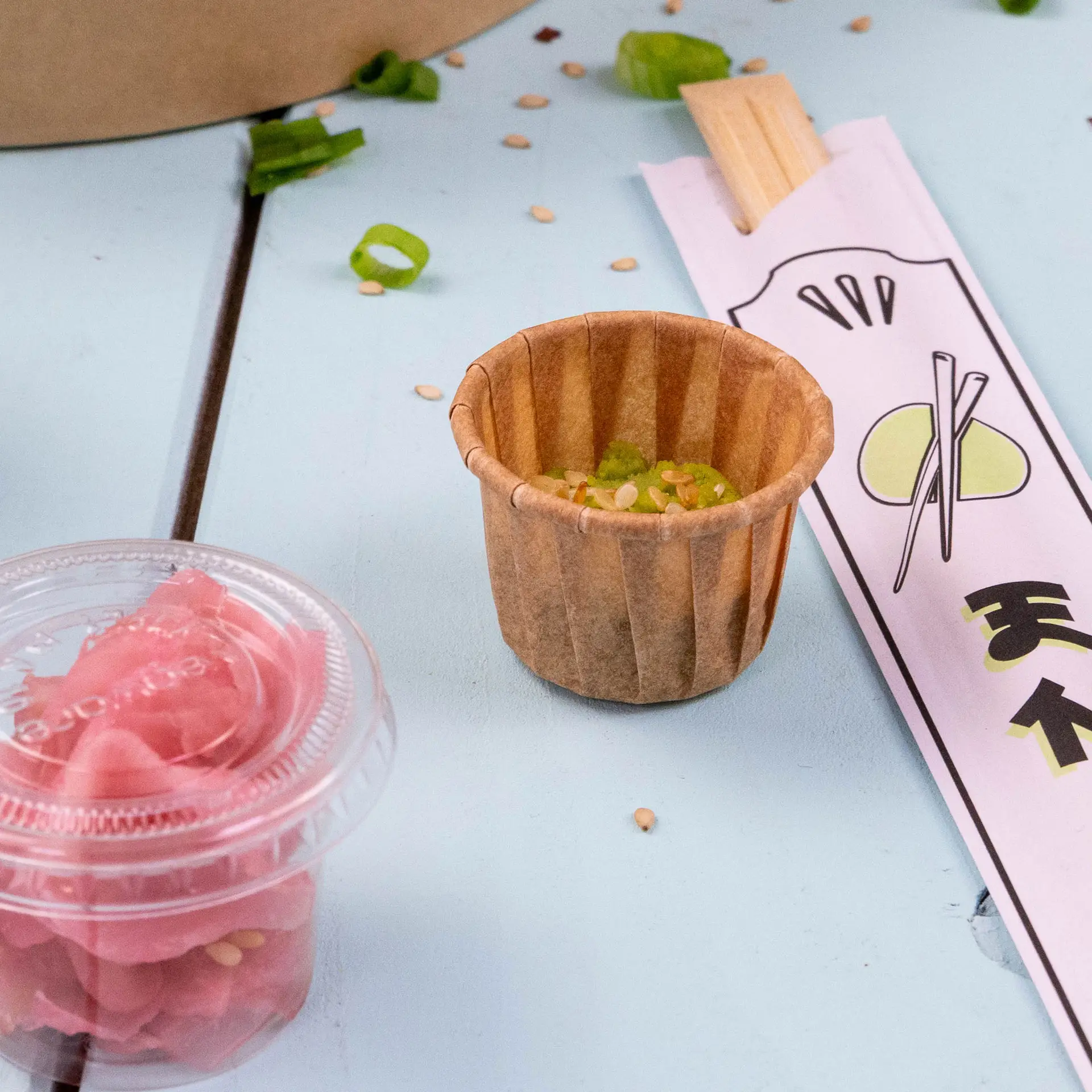 Brown paper cup for dips and sauces, 15 ml capacity, with sesame seeds and chopped green onions next to pickled ginger and chopsticks.