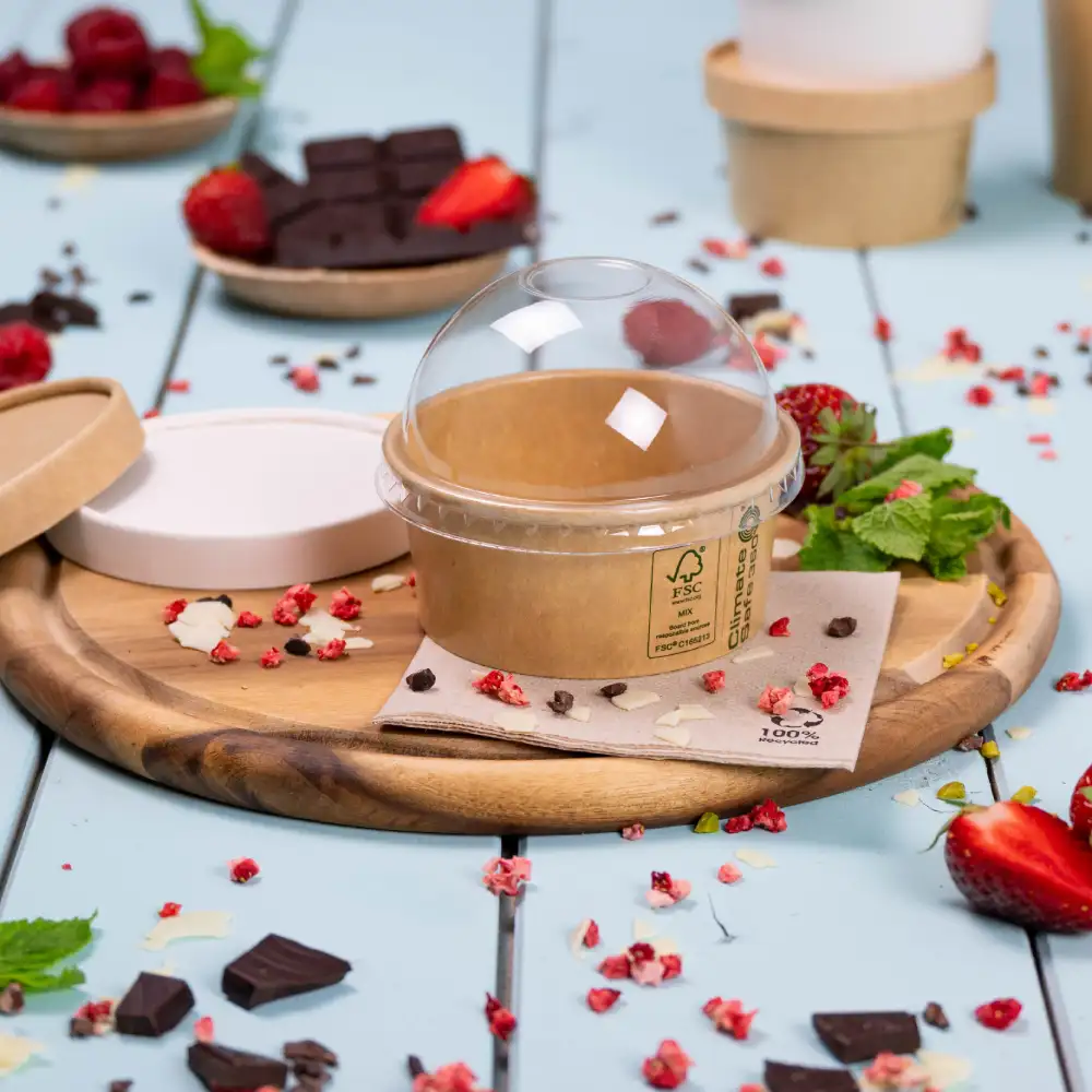 Takeaway dessert container with a transparent lid on a wooden tray, surrounded by fresh strawberries, raspberries, and chocolate pieces.