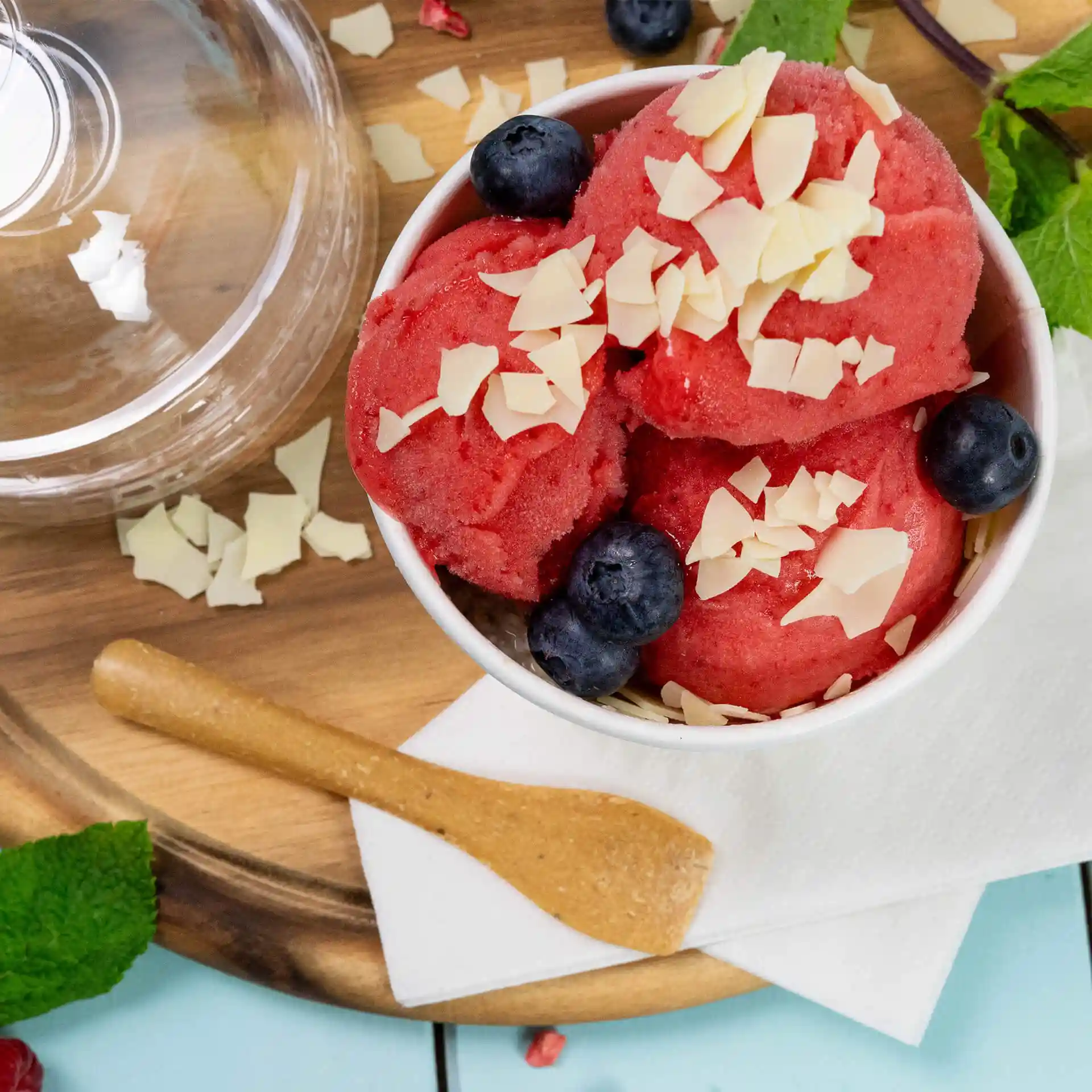 Schüssel mit rotem Sorbet, garniert mit Schokoladenraspeln und Heidelbeeren, begleitet von einem Holzlöffel auf einer Serviette.