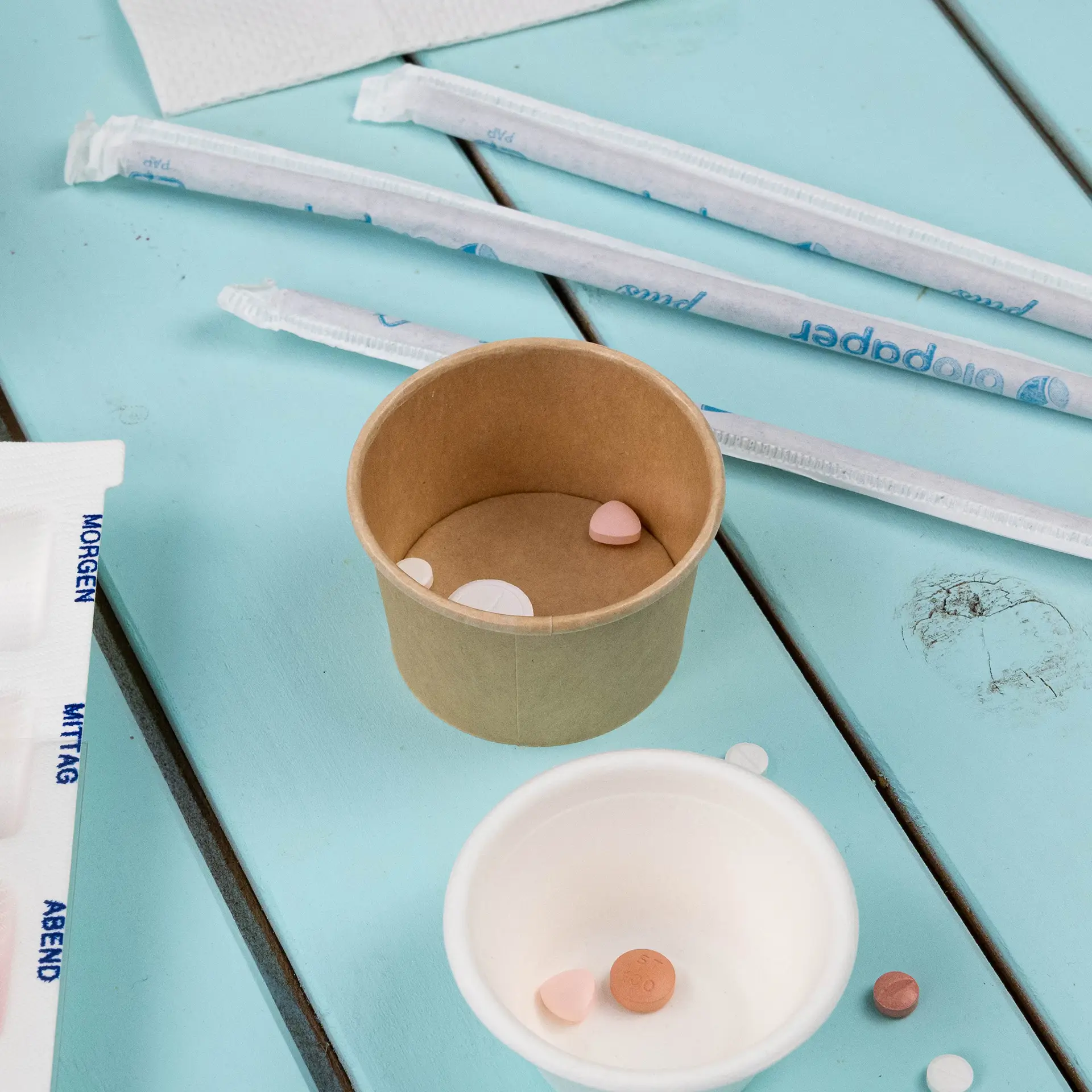 Ein ungebleichter Papp-Becher gefüllt mit kleinen, bunten Gegenständen; umgeben von einem weißen Becher und Papierstrohhalmen.