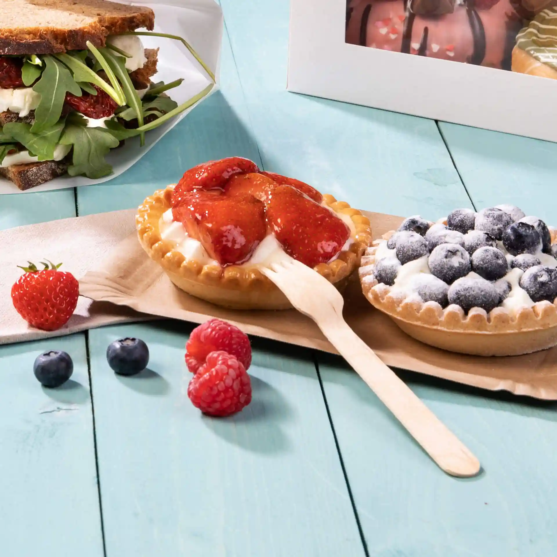 Bild von zwei Desserttörtchen mit Erdbeer- und Heidelbeerbelag, begleitet von frischen Beeren und einer Holzgabel auf einer hellblauen Oberfläche.