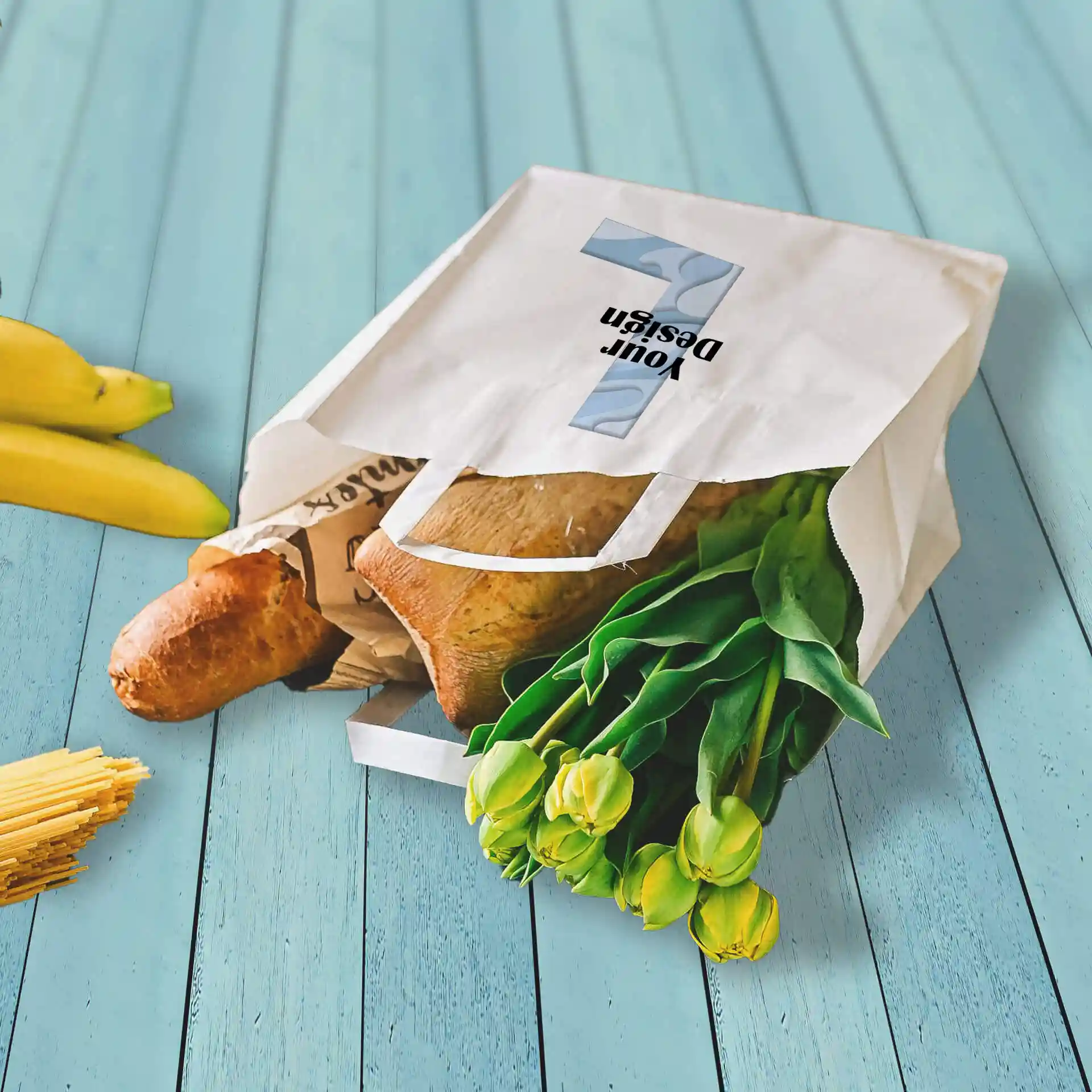 White paper carrier bag with groceries, including a baguette and tulips, on a blue wooden surface.