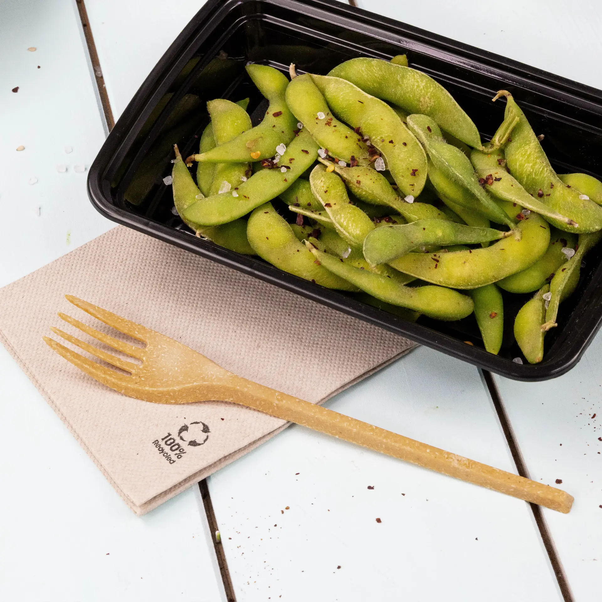 Bild von frischen Edamame-Bohnen in einem schwarzen Behälter, begleitet von einer biologisch abbaubaren Gabel auf einer recycelten Papierserviette.