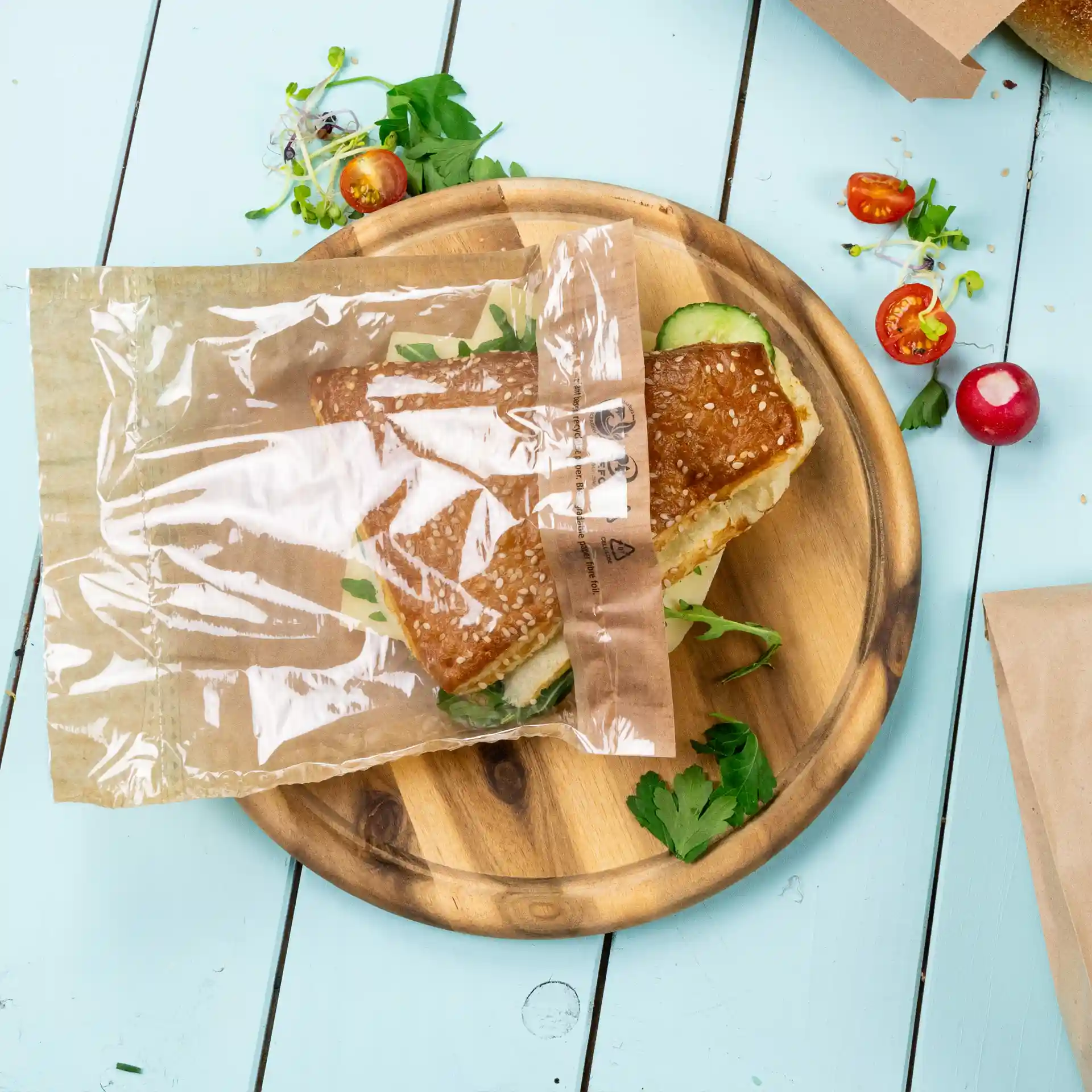 Transparente Snacktüte mit einem Sandwich darin, auf einem Holzbrett mit frischen Kräutern und Gemüse.