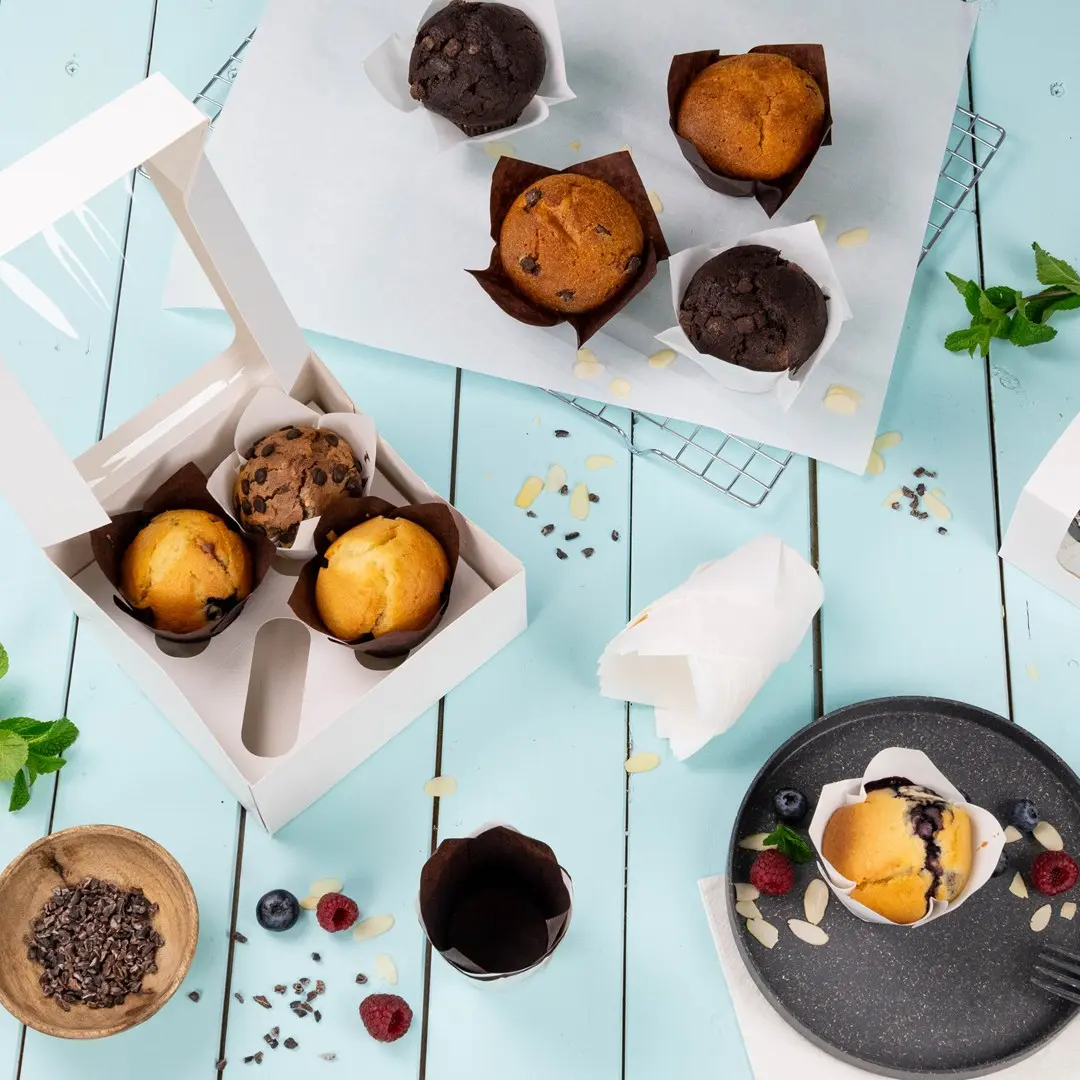 A white cupcake box containing three muffins, surrounded by additional muffins, berries, and chocolate pieces on a blue surface.
