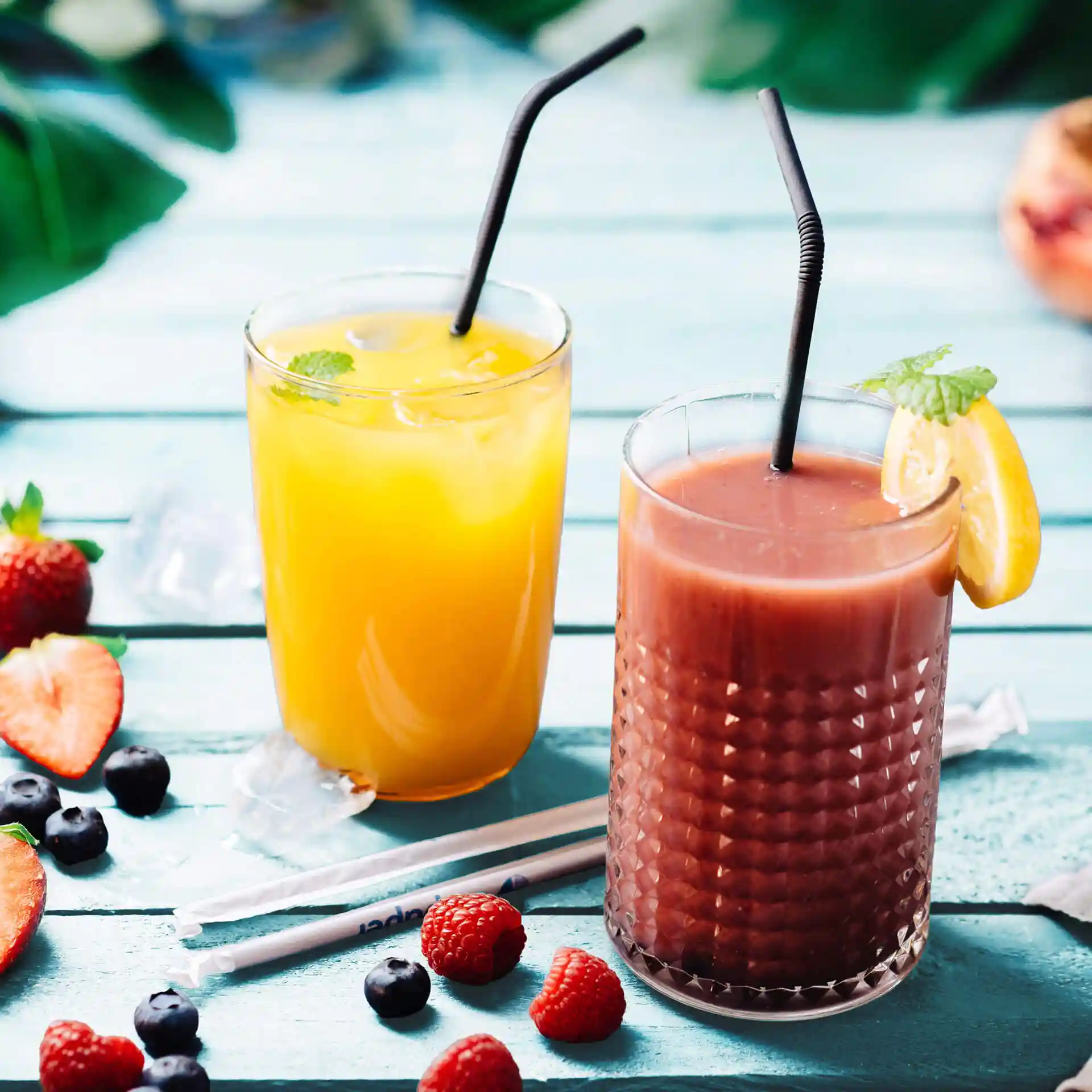 Bild von zwei Getränken in Gläsern mit schwarzen Strohhalmen, umgeben von Erdbeeren, Heidelbeeren und Himbeeren auf einer blauen Oberfläche.