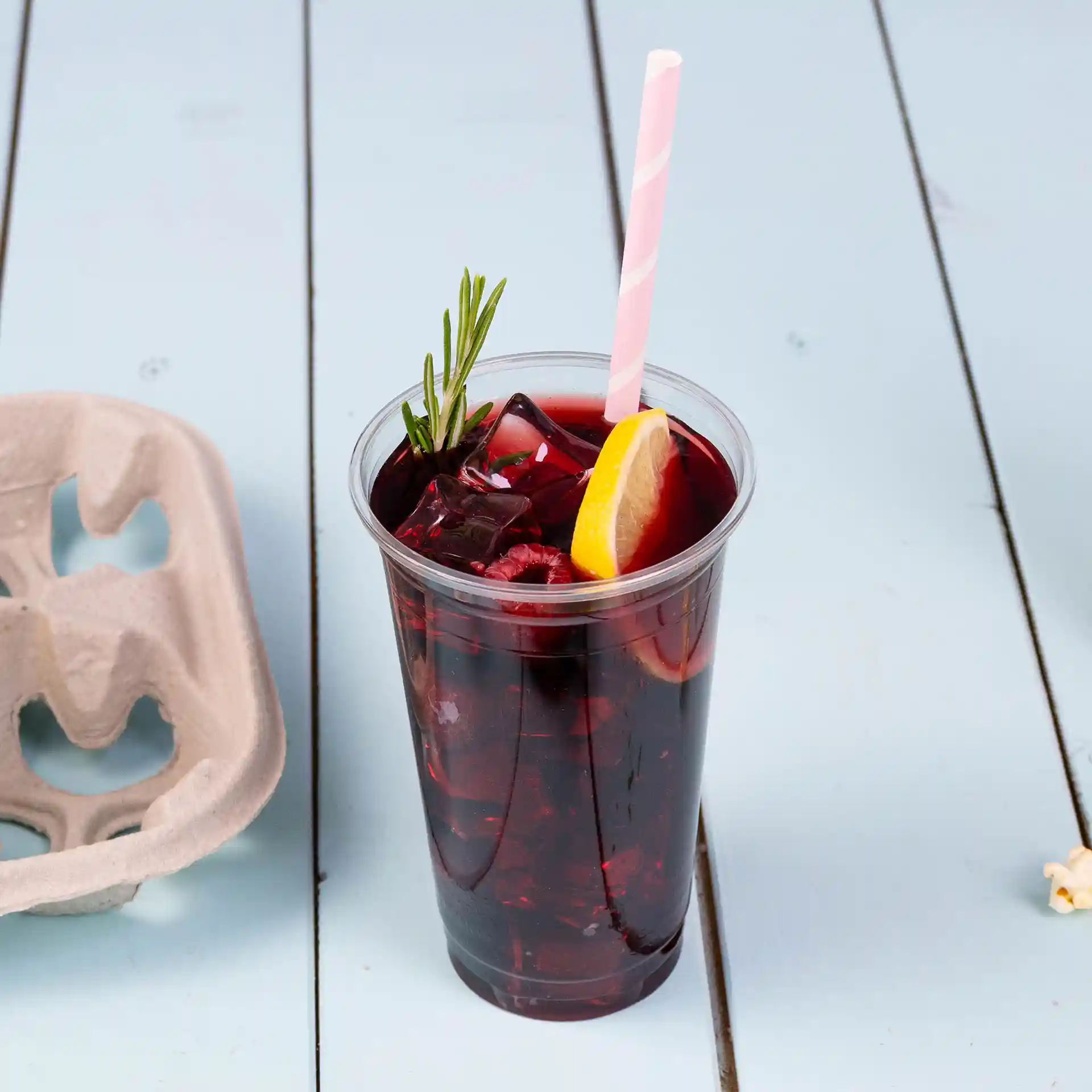 Klarer Plastikbecher gefüllt mit dunklem Getränk, Eis, Zitronenscheibe und Rosmarinzweig, mit einem pink gestreiften Strohhalm auf einer hellblauen Oberfläche.