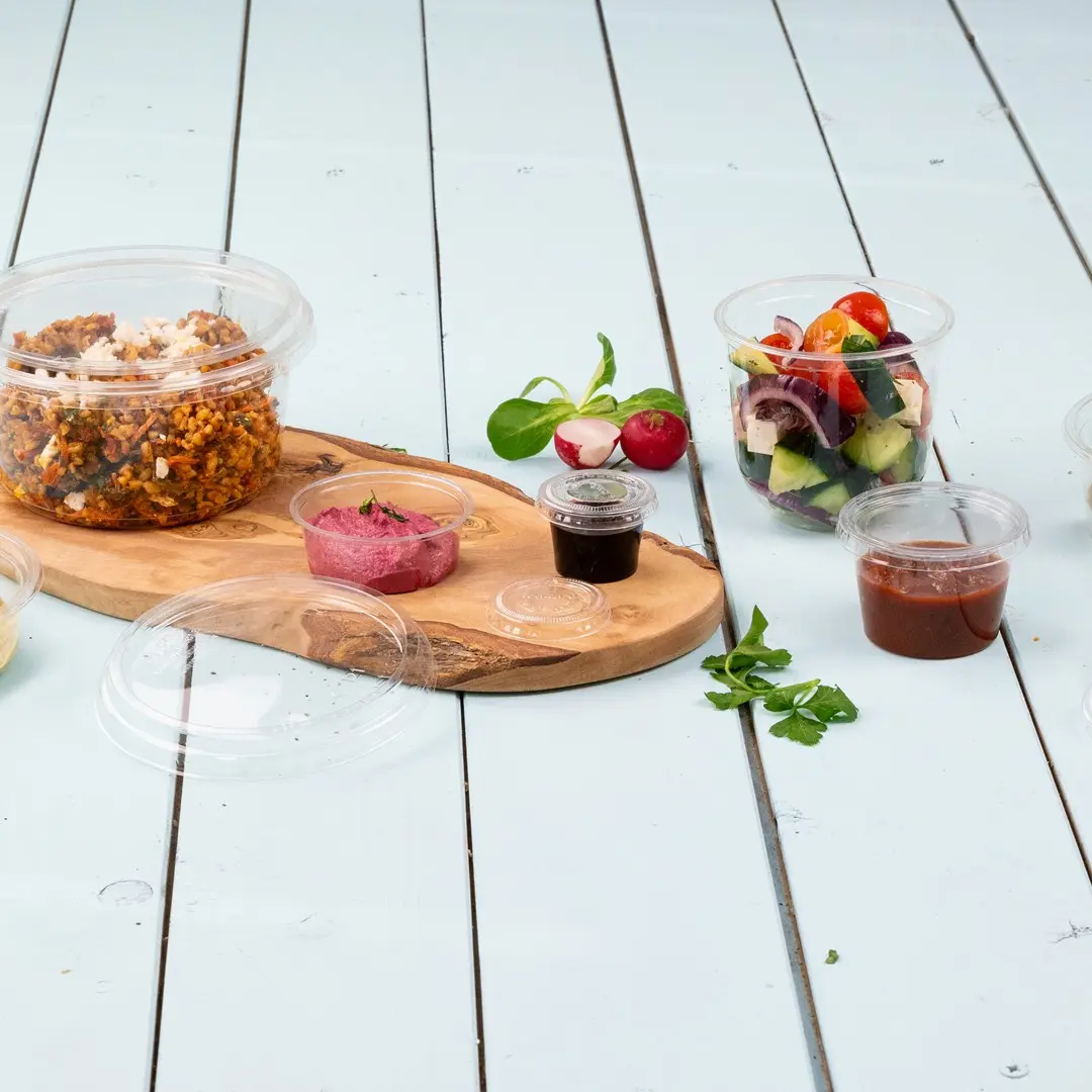 Klare Kunststoffbehälter mit verschiedenen Lebensmitteln, darunter ein Salat, Dips und ein Getreidegericht, auf einem Holzbrett angeordnet.