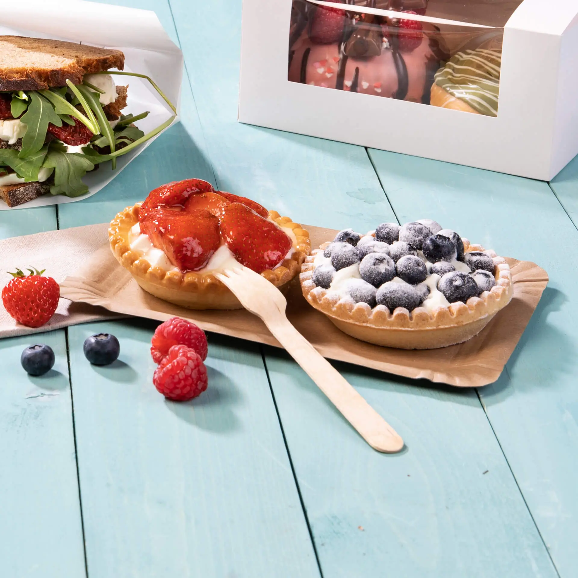 Zwei Desserttorten mit Erdbeeren und Heidelbeeren auf einem braunen Pappgeschirr, daneben frische Beeren und ein Sandwich.