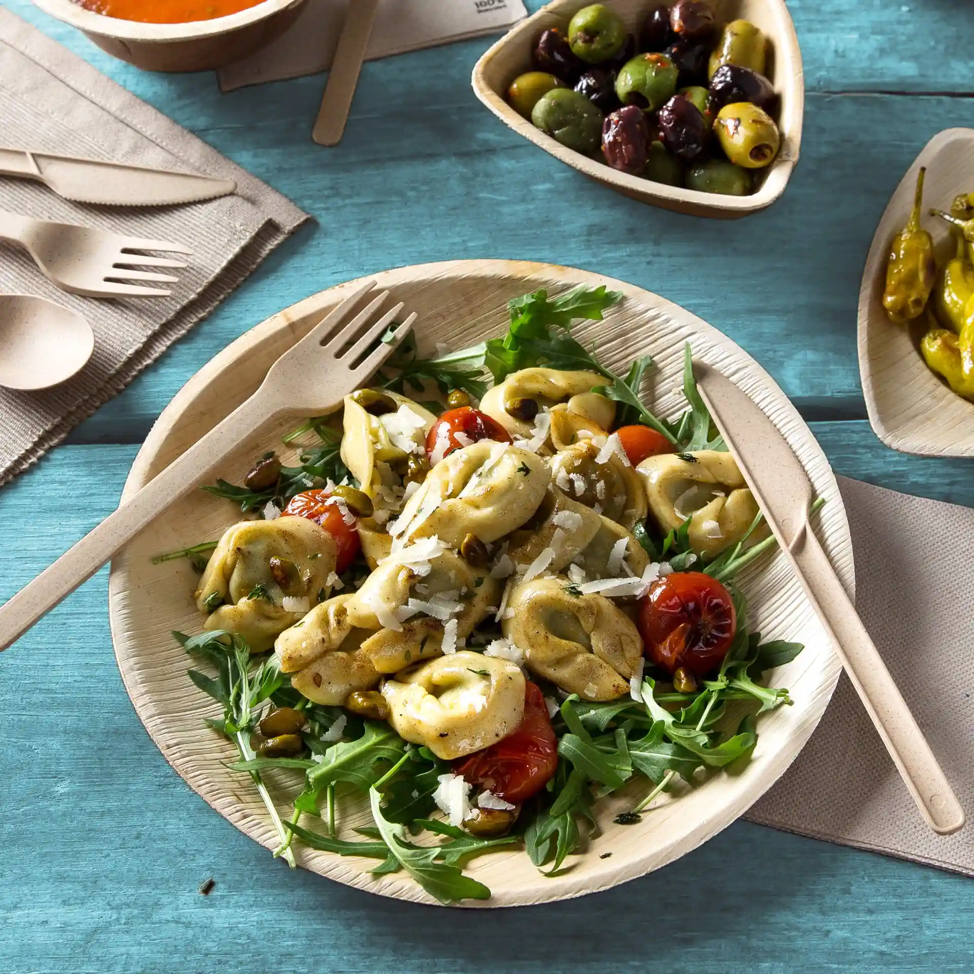 Runde Palmblattplatte mit Salat, Tortellini und Kirschtomaten, begleitet von Holzutensilien auf einer blauen Oberfläche.