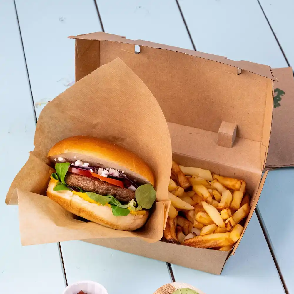 Burger in einer Papierverpackung mit frischem Gemüse neben einer Box Pommes auf einer blauen Oberfläche.