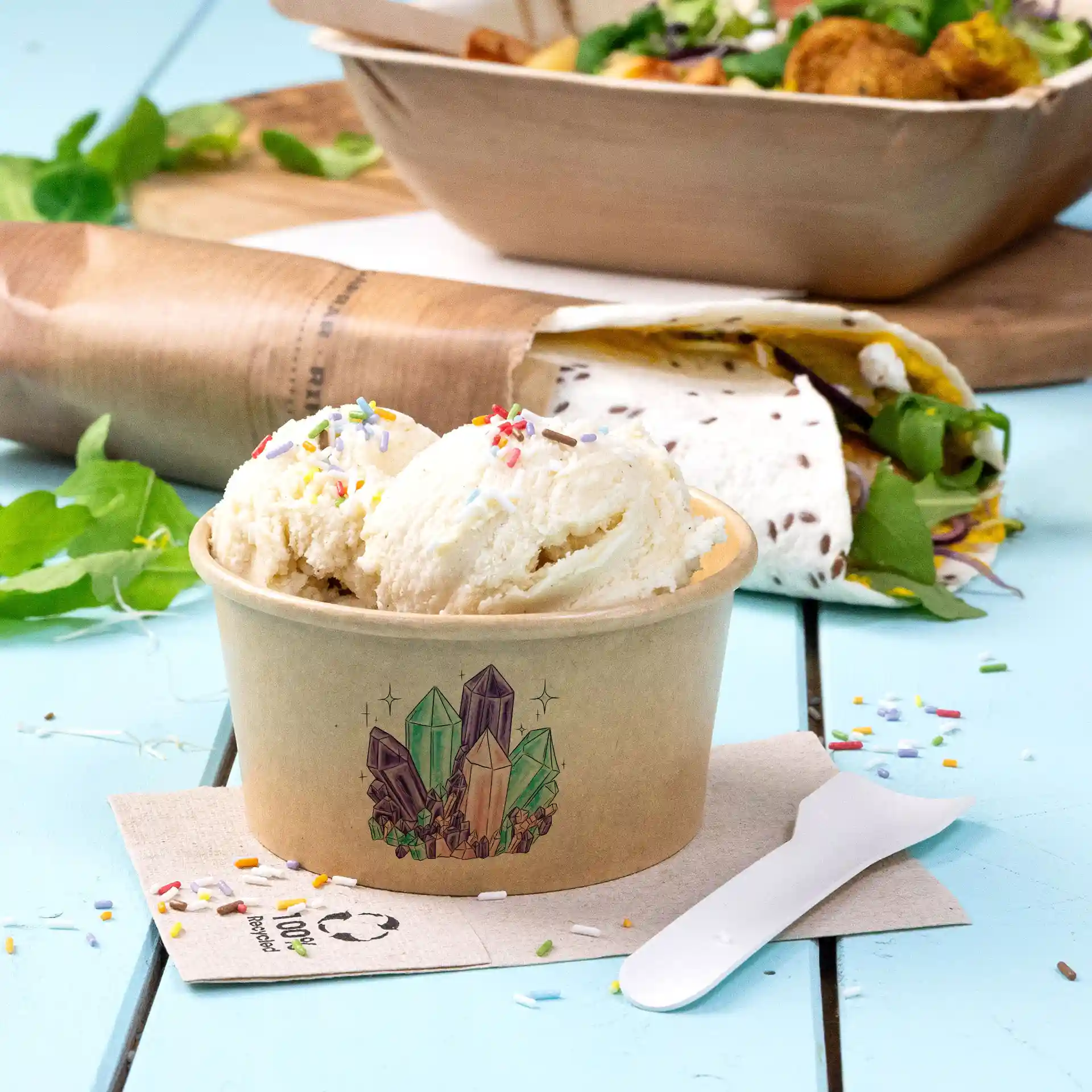 Runder brauner Becher mit drei Kugeln Eis und bunten Streuseln, auf einer blauen Oberfläche platziert.