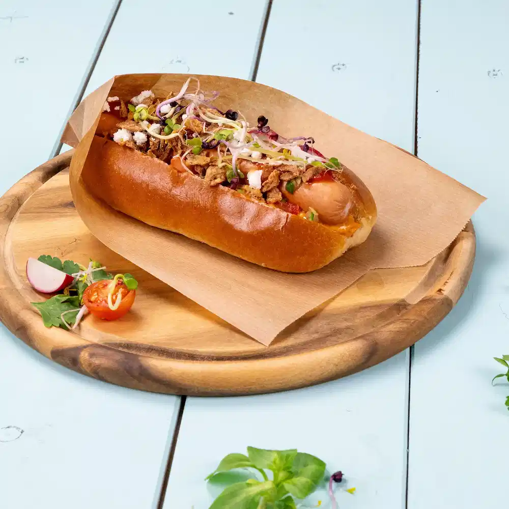 Hot Dog in weichem Brötchen, belegt mit knusprigen Zwiebeln, Sprossen und garniert mit Kirschtomaten und Rettich auf einem Holzbrett.