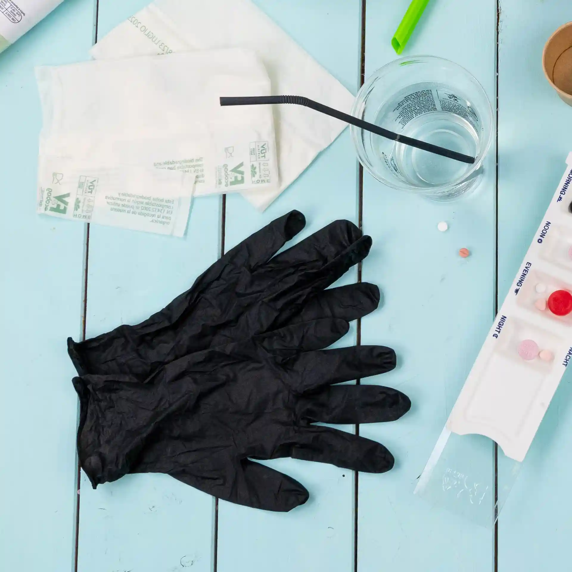 Schwarze Nitrilhandschuhe in Größe M auf einer blauen Holzoberfläche mit einem Glas Wasser, einem Strohhalm und anderen Gegenständen.