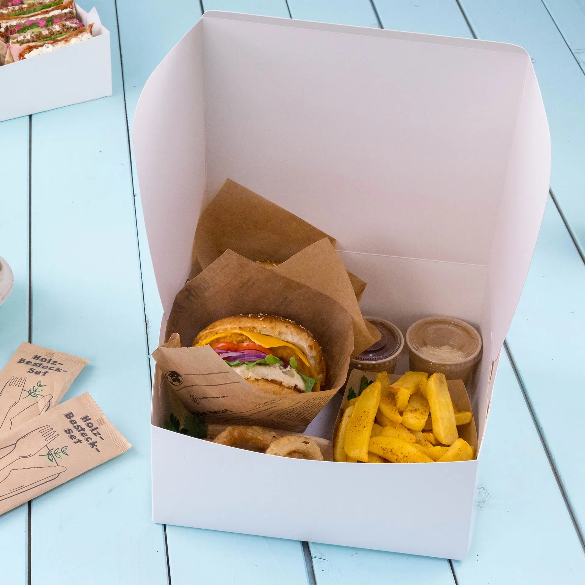 Takeaway box containing a burger with toppings, fries, and two small containers, all on a light blue surface.