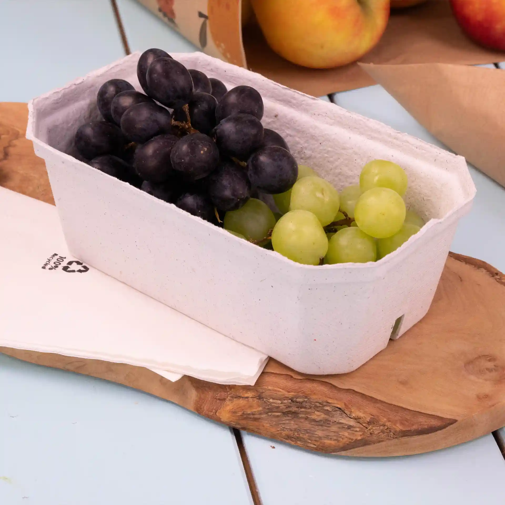 Container with black and green grapes inside a white wood pulp punnet on a wooden surface with napkins nearby.