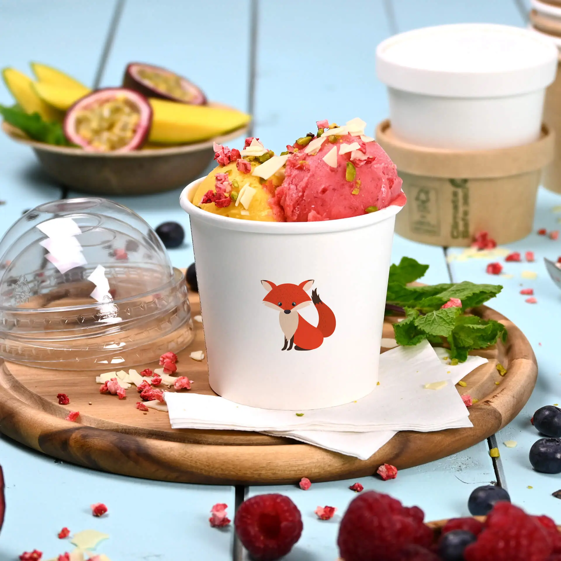 White ice cream cup featuring a fox illustration, filled with pink and yellow ice cream and garnished, on a wooden board.